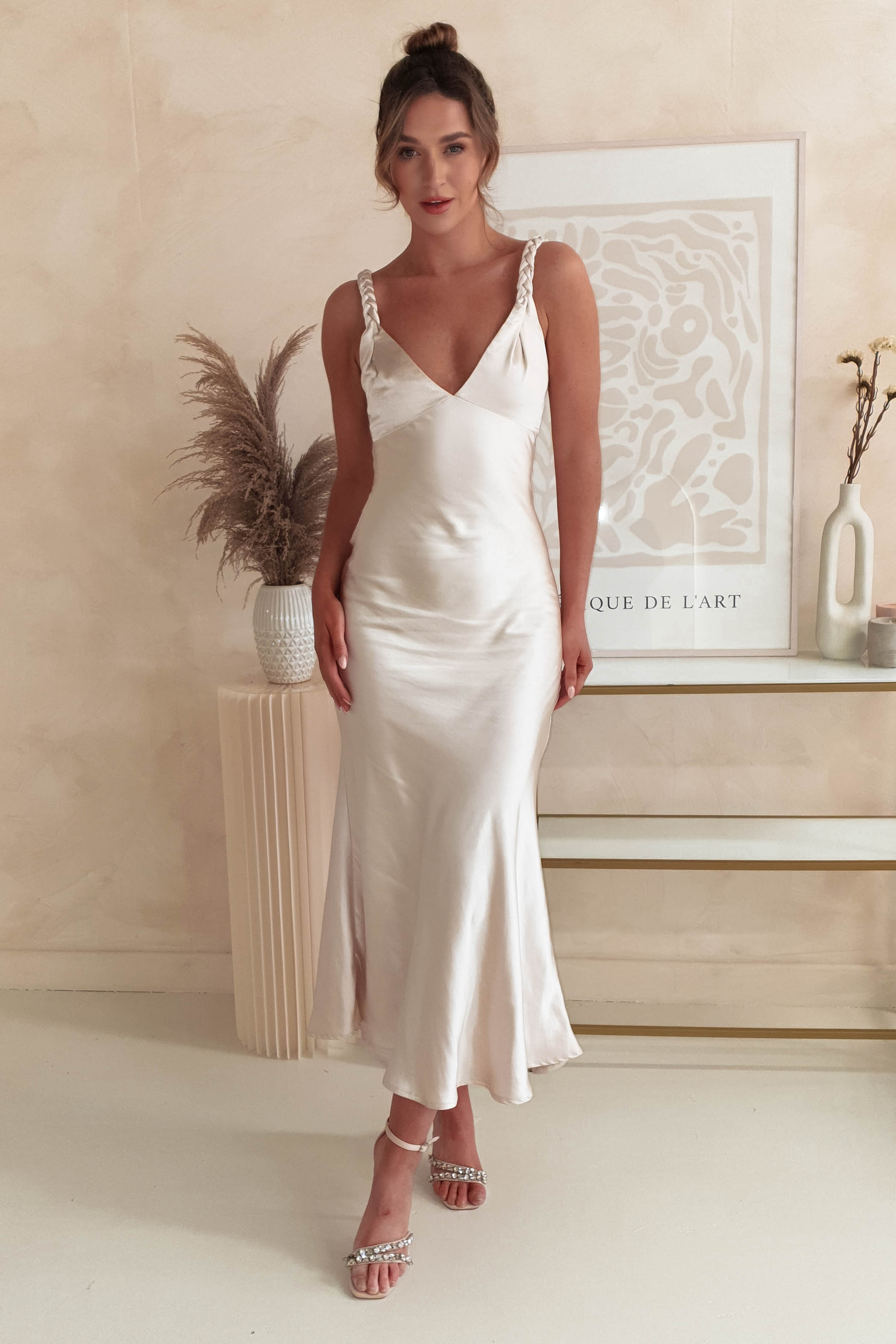 A woman in a satin maxi dress with plaited straps and strappy heels stands elegantly in front of modern art and decor, ready for special occasions.