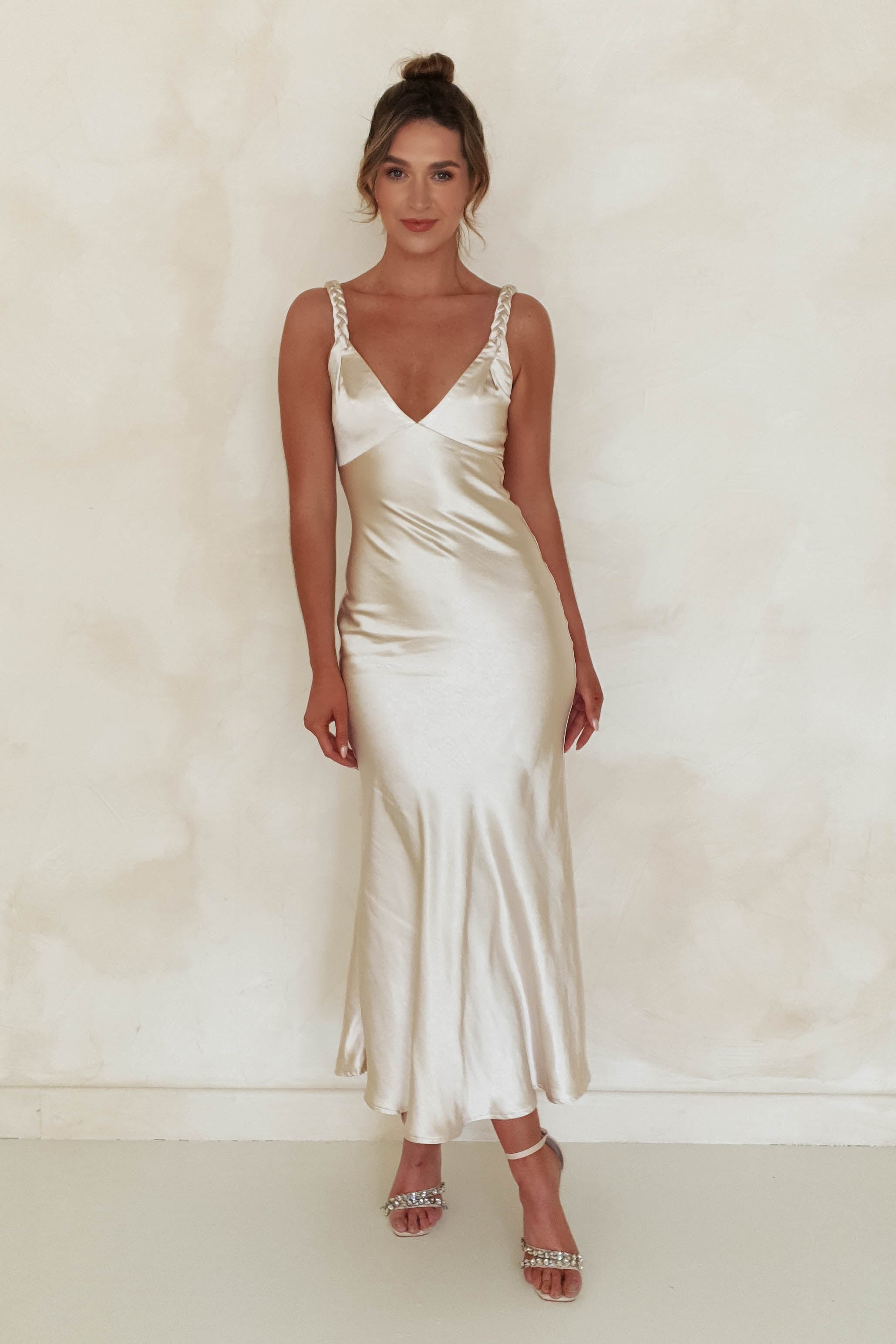 A woman in a sleek satin maxi dress with jeweled, plaited straps stands against a soft, textured background. Perfect for special occasions, she completes the look with strappy heels and her hair styled in a neat bun.