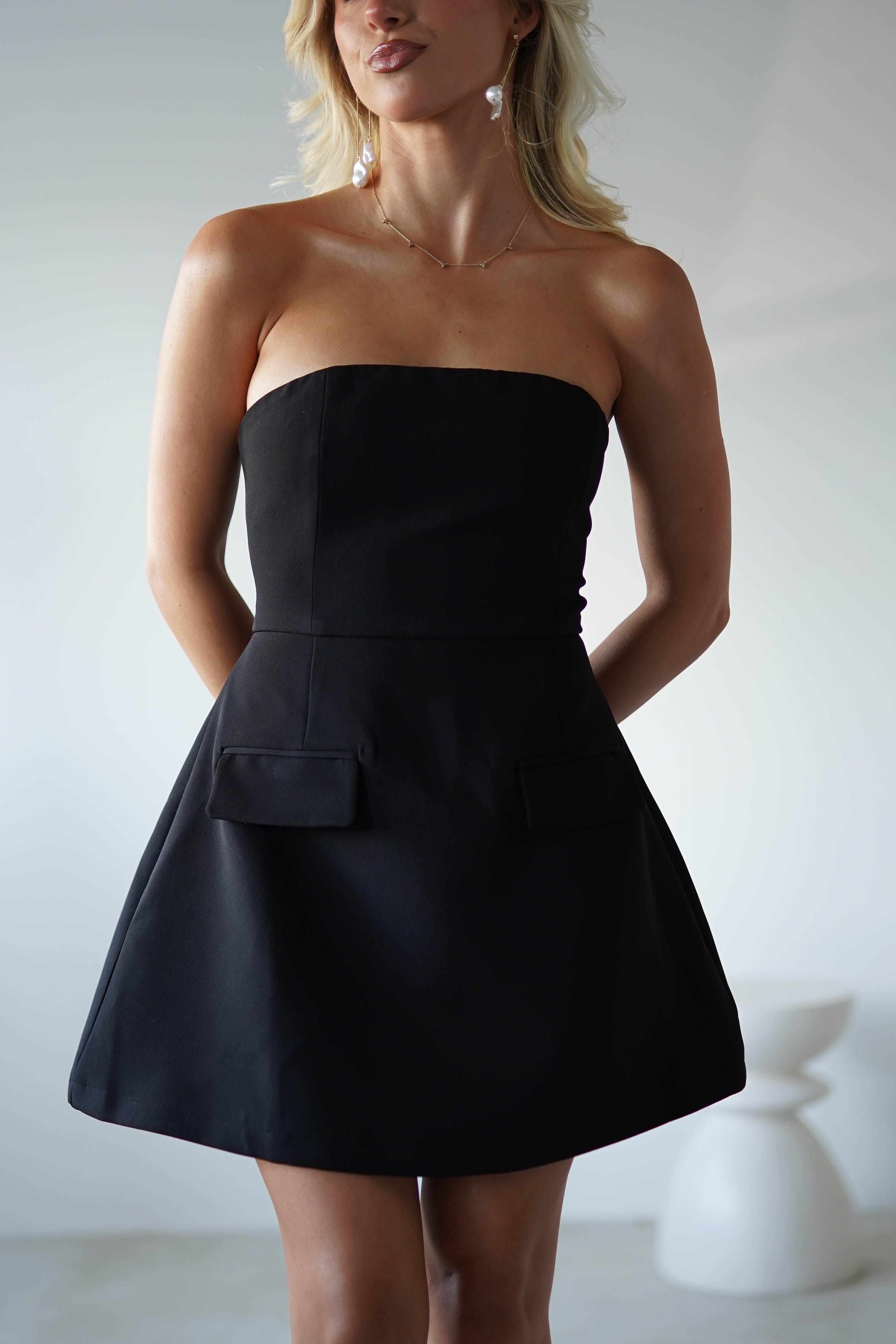 A woman wearing a strapless mini dress stands indoors with her hands behind her back. The polyester dress features a fitted bodice and a flared skirt with two front pockets—perfect for a stylish wedding guest. She wears earrings and a delicate necklace as the background softly blurs.