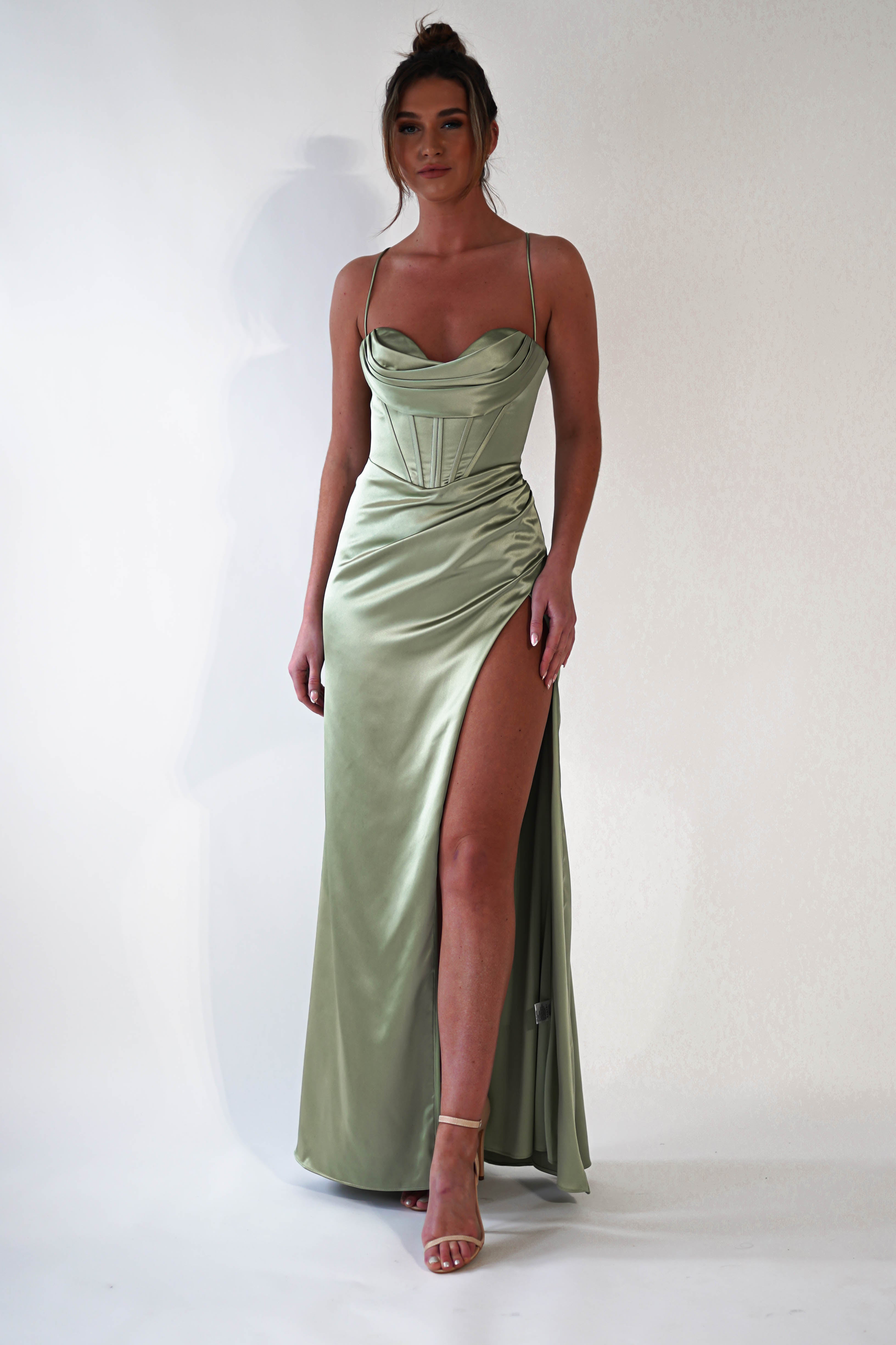 A woman poses in a light green satin corset dress with thin straps and a high thigh slit—perfect for prom or any special occasion. She wears nude heels and stands against a plain white background.