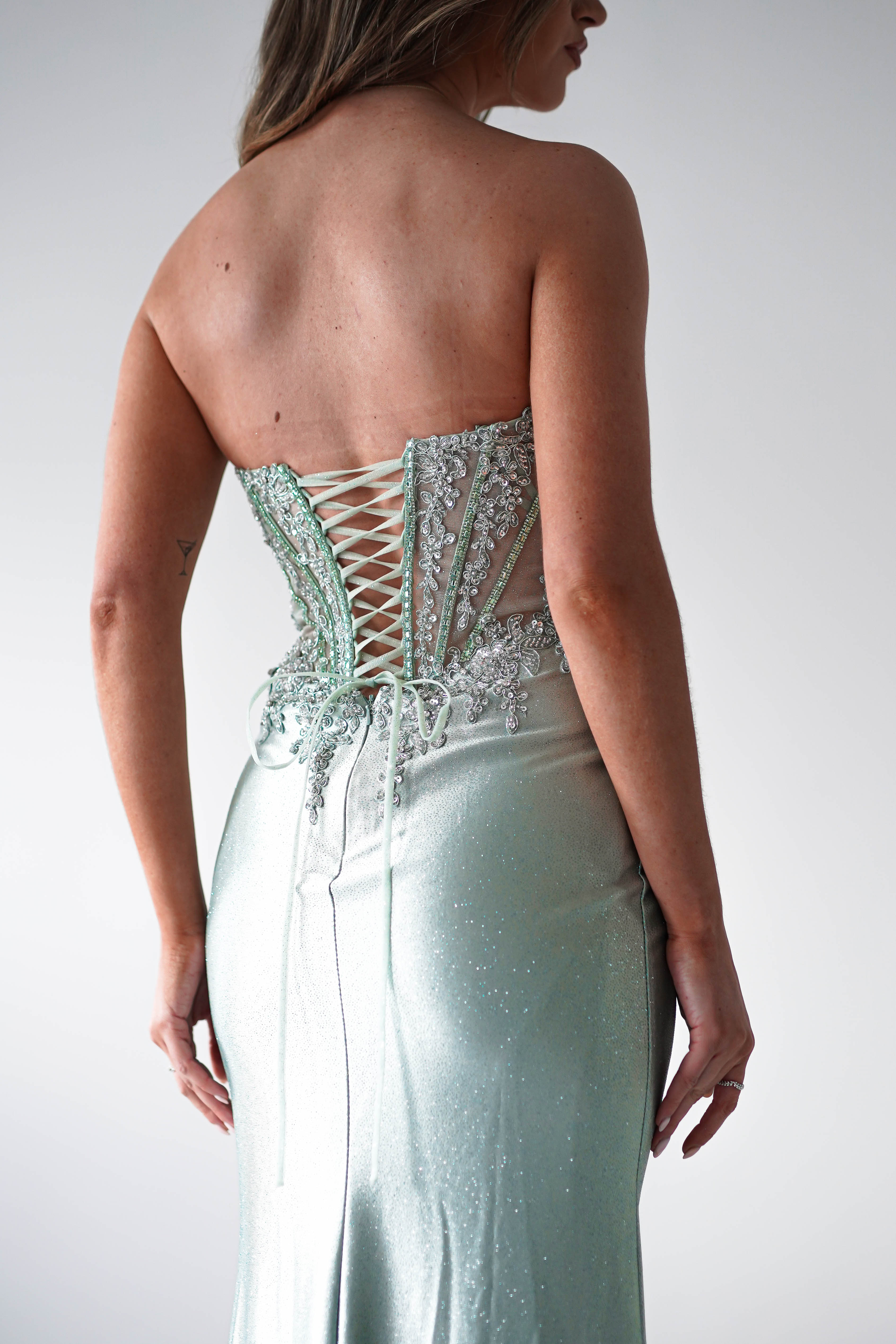 A person wearing a strapless, sequin-embellished gown with intricate lace-up detailing and bead embellishments on the back. Perfect for formal occasions, they stand against a plain background, showcasing the elegant design and smooth fabric.