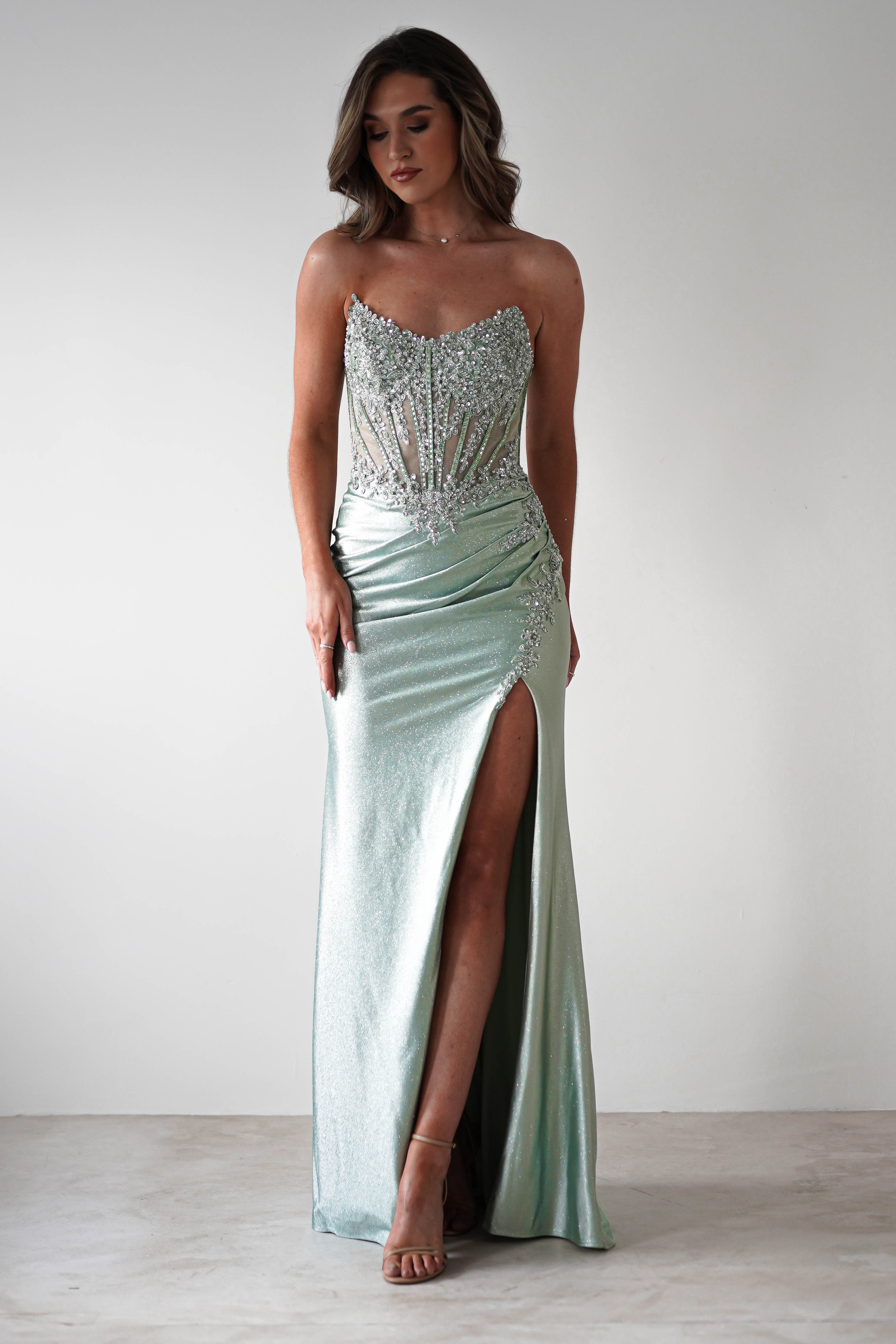 A woman stands wearing a shimmering, strapless mint green gown with an intricate lace bodice and corset midriff. She poses confidently, her long wavy hair cascading down as she pairs the ensemble with nude heels, ready for formal occasions against a plain backdrop.