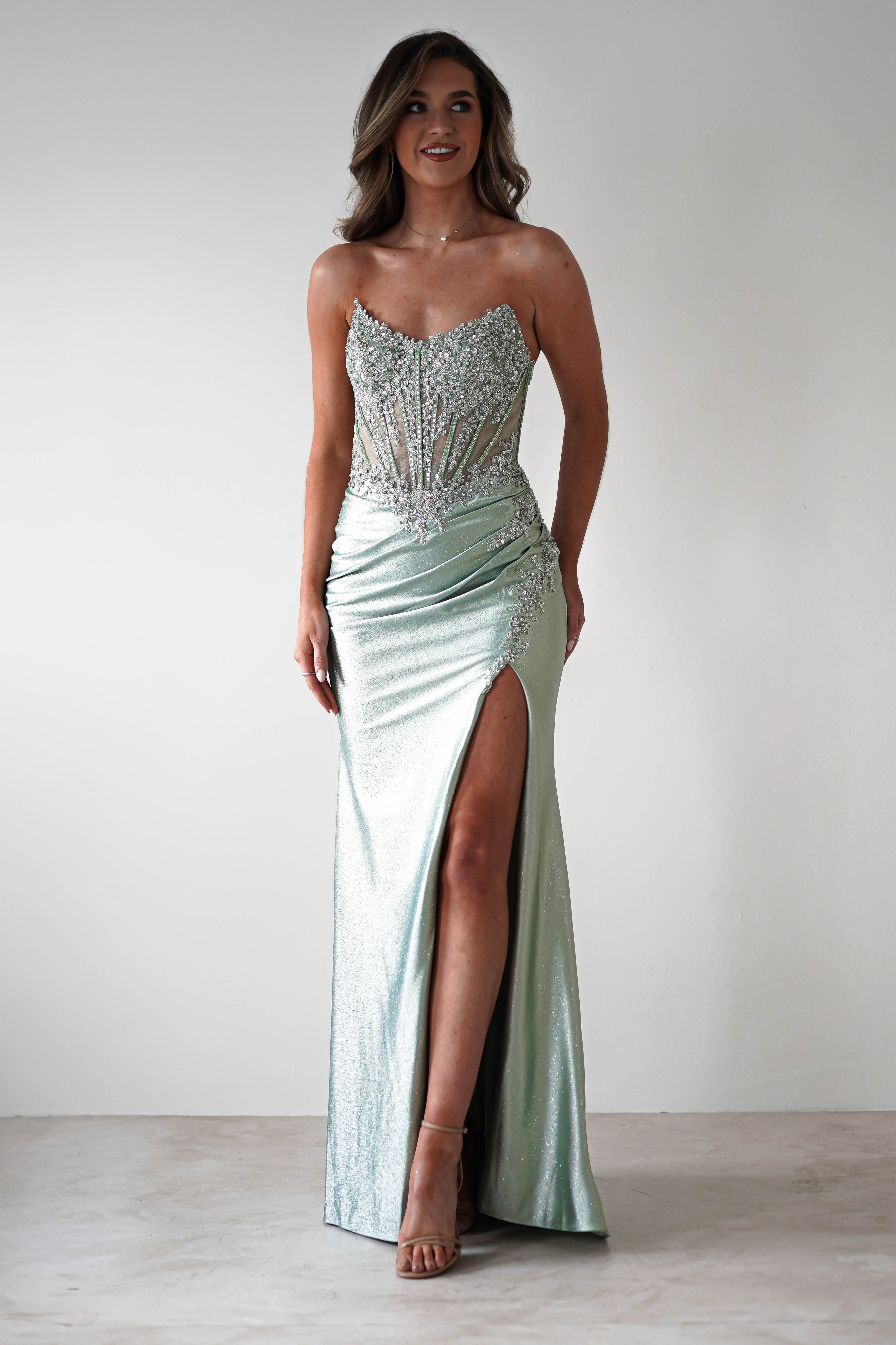 A woman in a strapless, sequin-embellished light green gown with a high slit and intricate beadwork gracing the corset midriff. She stands on a plain white background, wearing high heels, her hair styled in loose waves, perfectly suited for formal occasions.