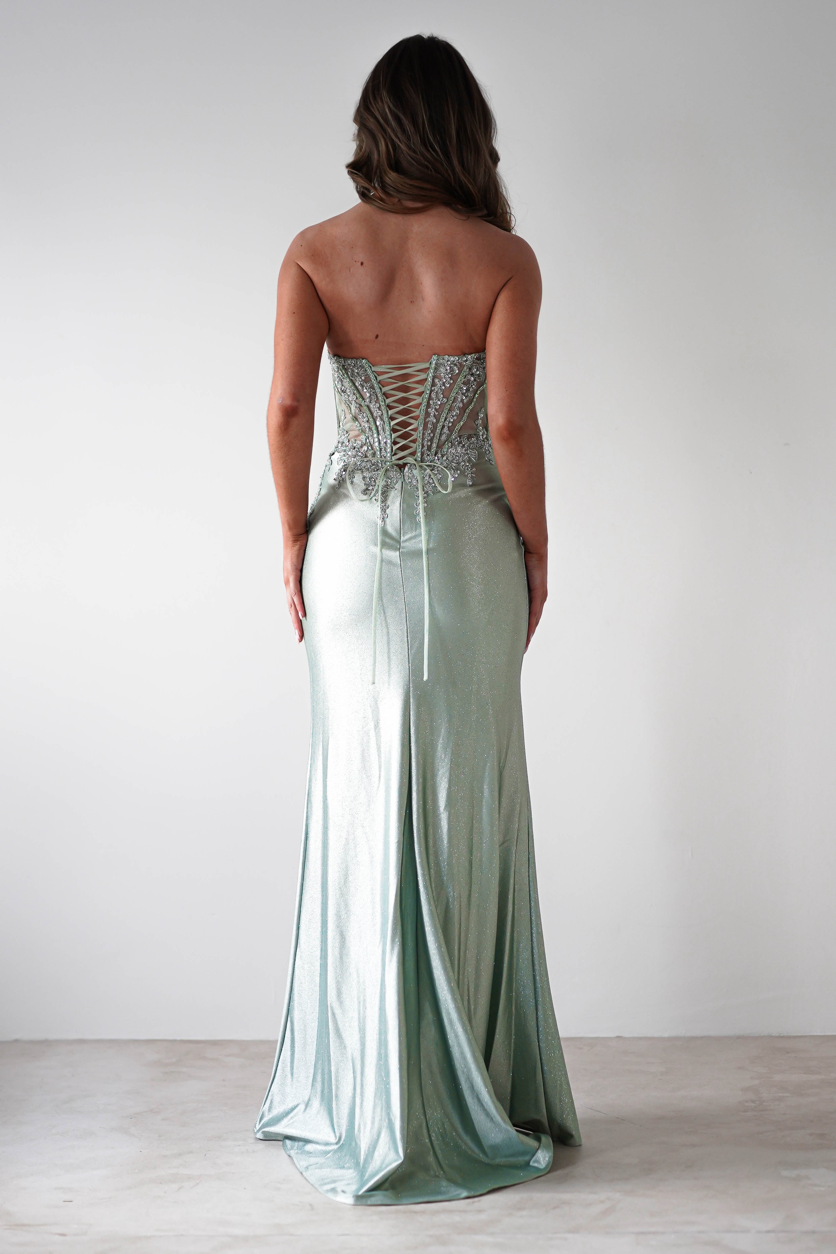 A woman in a strapless, sequin-embellished gown stands facing away, showcasing the lace-up back design. Perfect for formal occasions, this shimmering silver-green dress gracefully flows to the floor against a plain, light background.