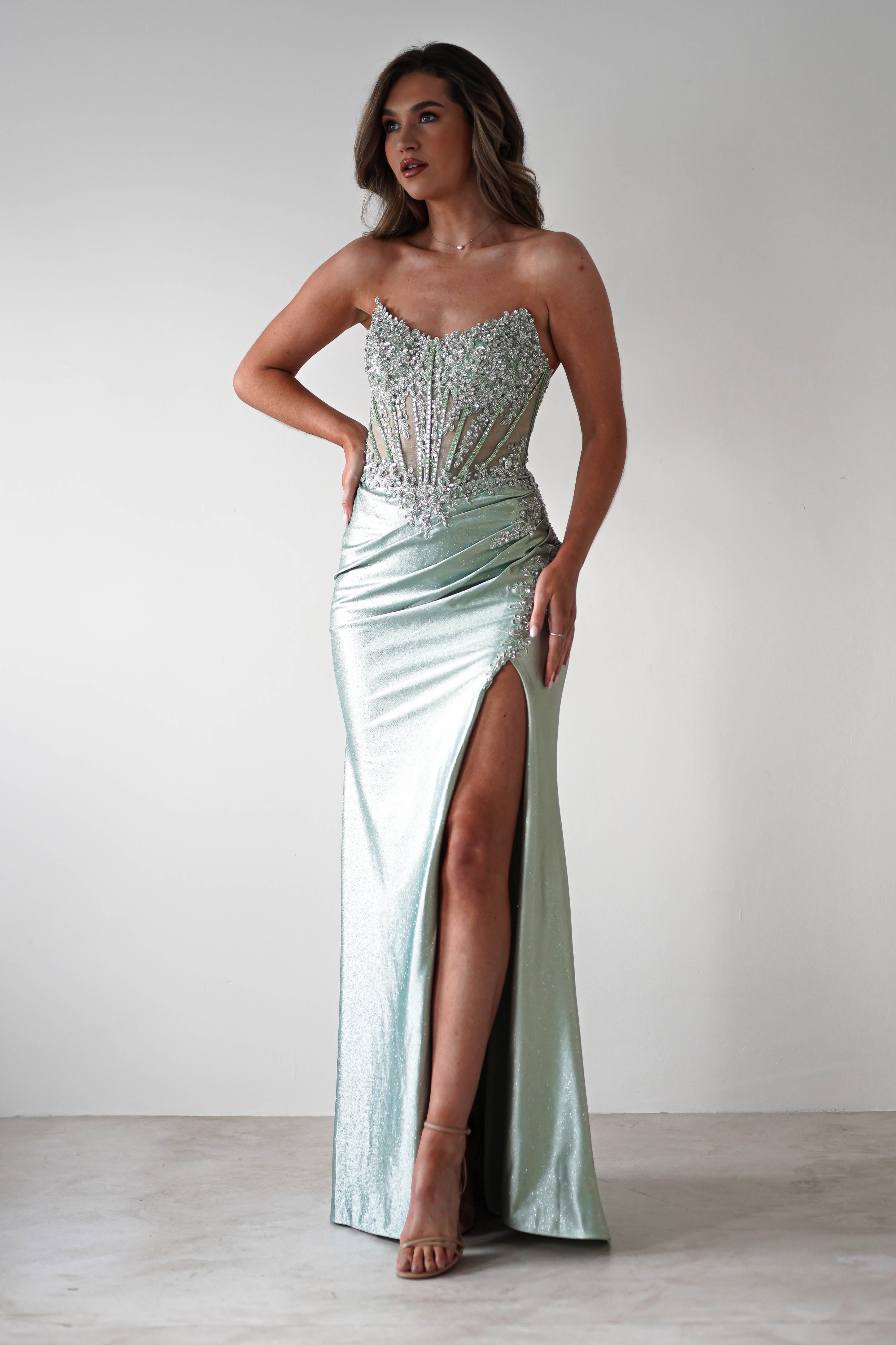 A woman poses in a sequin-embellished gown, perfect for formal occasions. The elegant light green dress features a strapless, beaded bodice and corset midriff with a high slit on one side. She stands on high heels against a neutral backdrop, one hand on her hip and the other by her side.