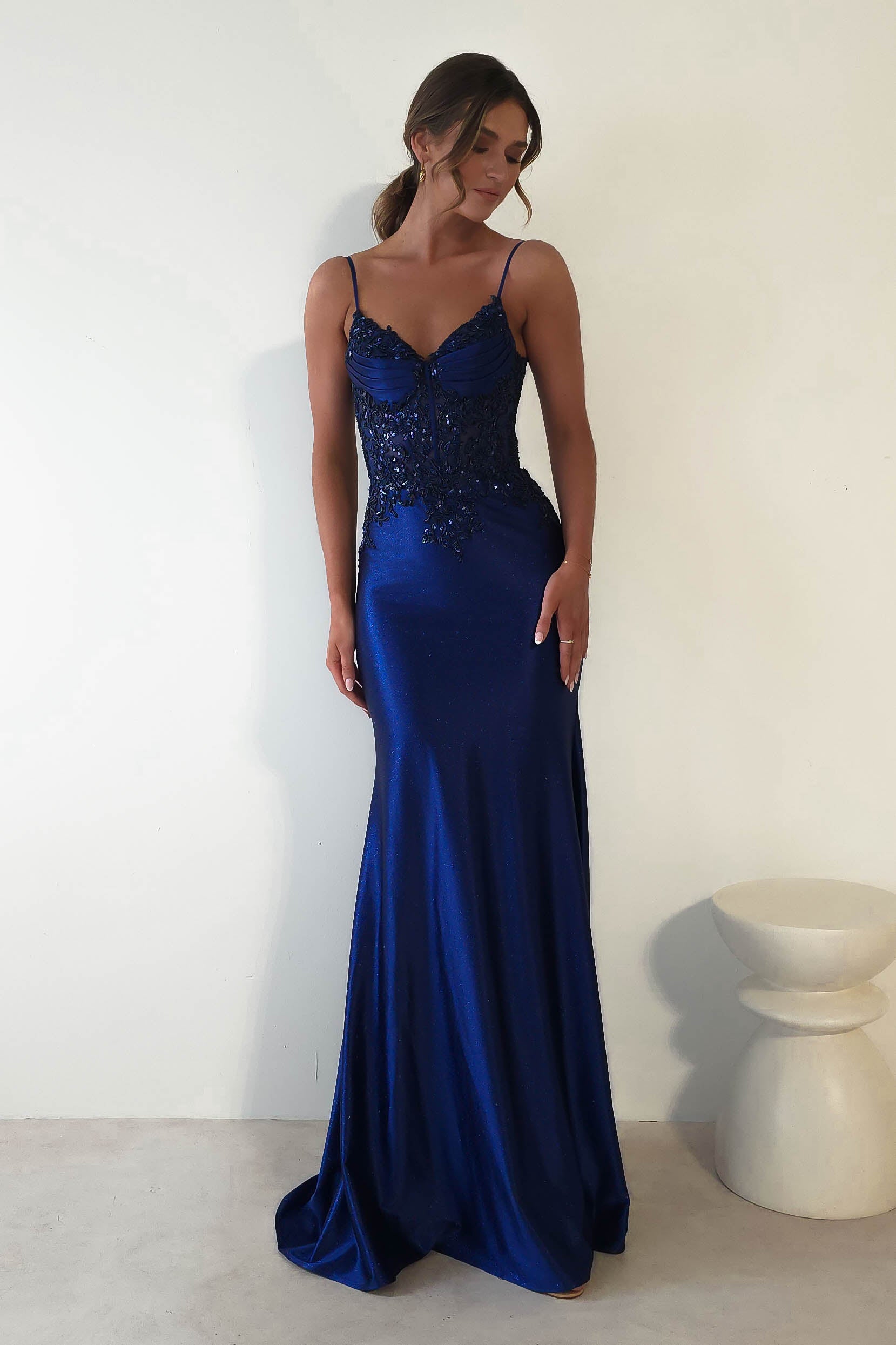 A woman in a mermaid glitter gown with intricate lace detailing stands beside a simple white stool against a plain background. She is looking down, highlighting the embellished bust and floor-length silhouette with spaghetti straps, perfect for formal events.