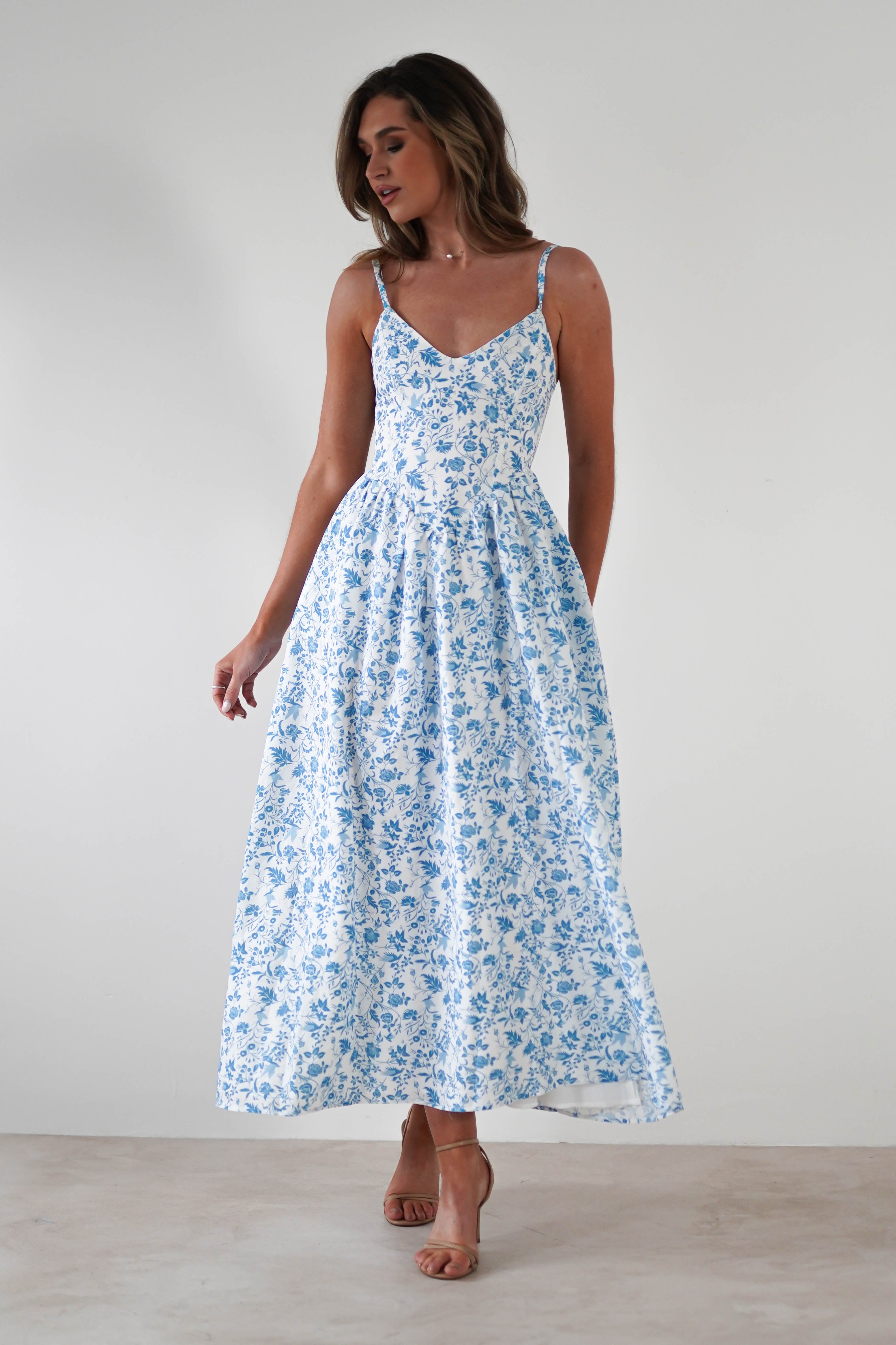 Woman wearing a summer print maxi dress with blue floral patterns and adjustable ties, standing against a plain light background, her head turned slightly to the side and one hand holding her skirt—perfect for wedding guests.