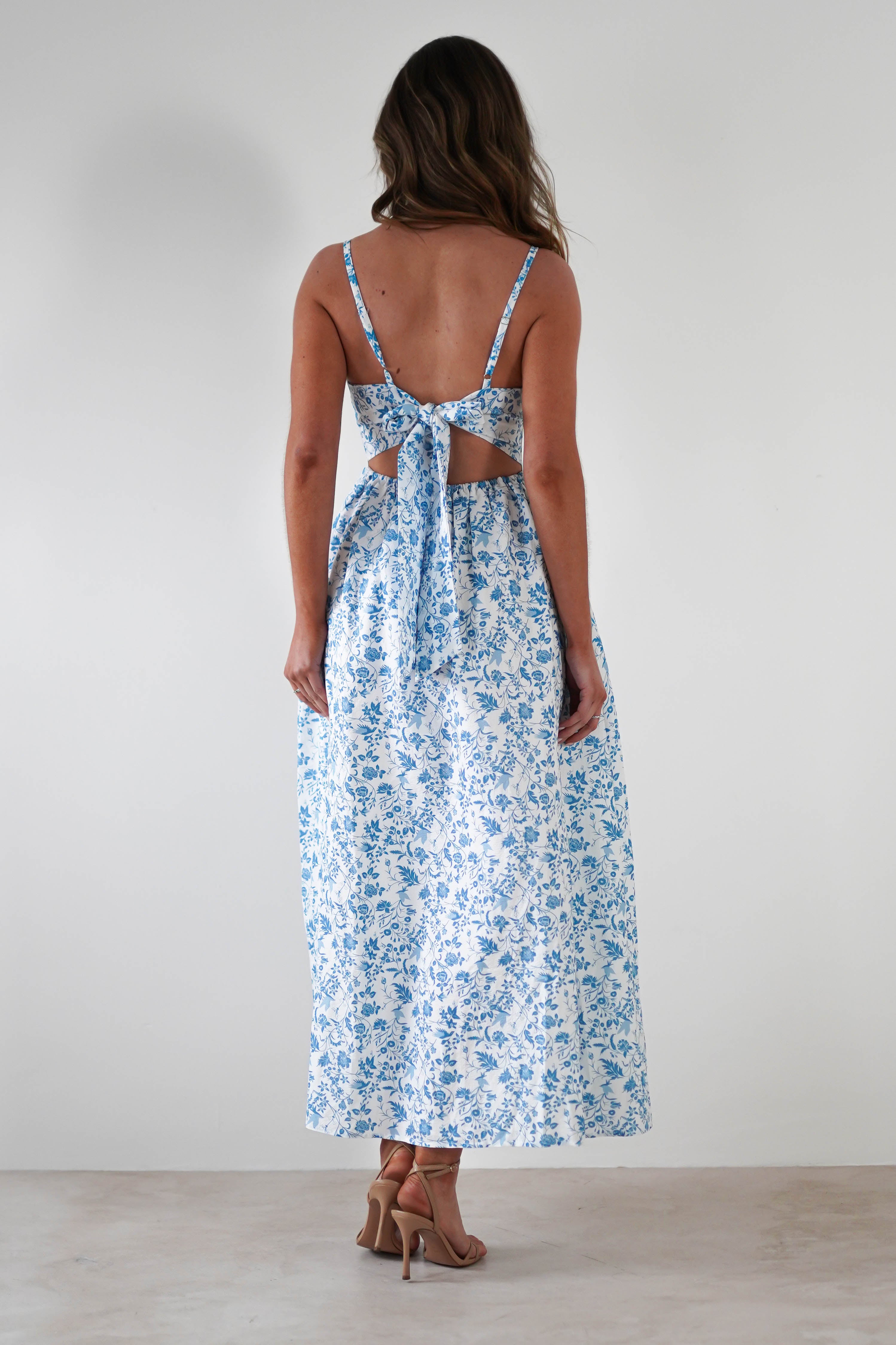 A woman stands facing away in a summer print maxi dress with blue florals, open back, large bow tie, and adjustable ties. Her loose hair flows as she wears tan high-heeled sandals—perfect for wedding guests against a plain light background.