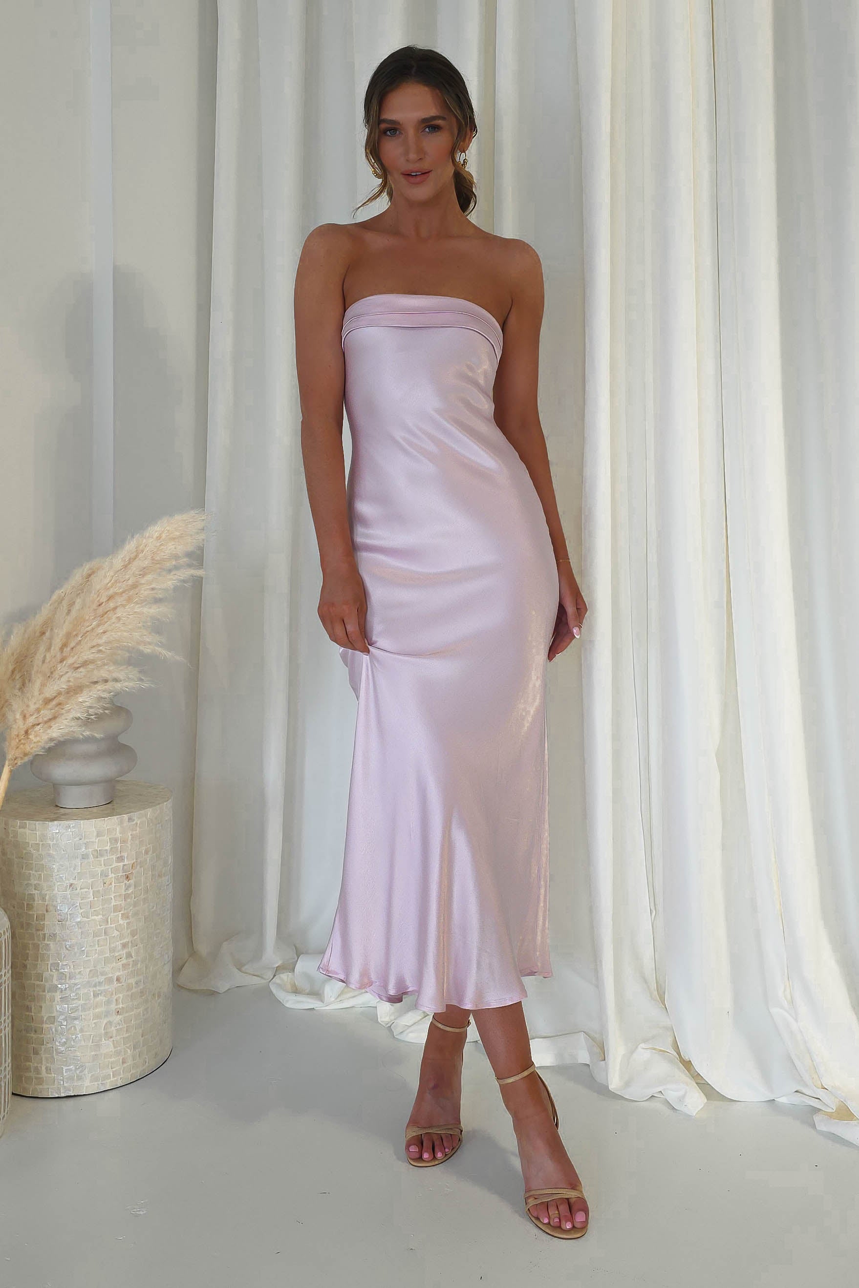 A person wearing a strapless midi dress in super soft, silky light pink fabric stands on a white floor against flowing white curtains. With long hair and strappy heels, they exude elegance. To the left, a decorative vase with pampas grass completes the chic ensemble perfect for wedding guests.