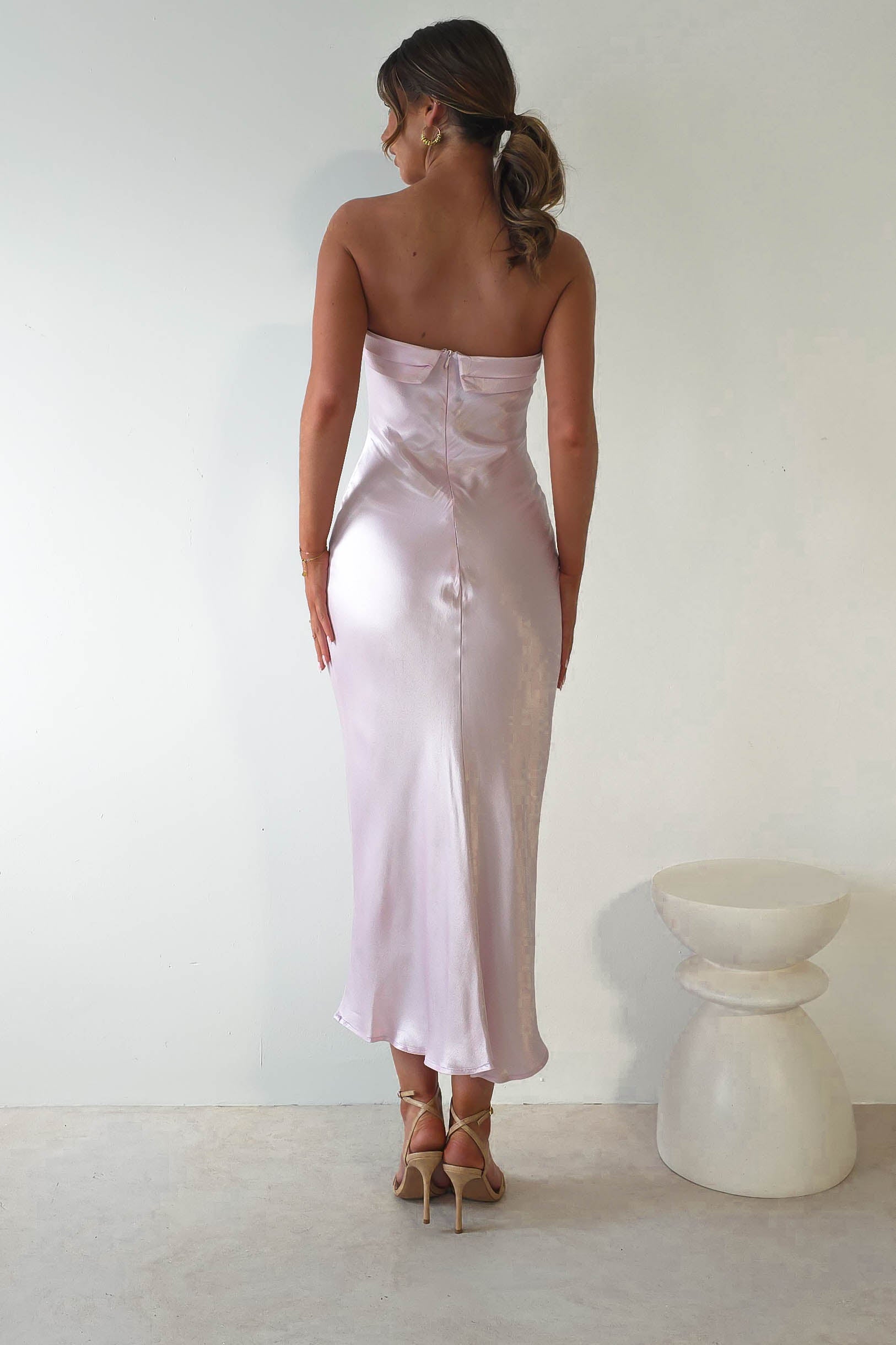 A woman with tied-back hair stands facing a wall, wearing a strapless midi dress in light pink, satin, and high heels. Perfect for wedding guests, the dresss super soft fabric drapes elegantly. The floor is light-colored, and a small white stool is next to her.