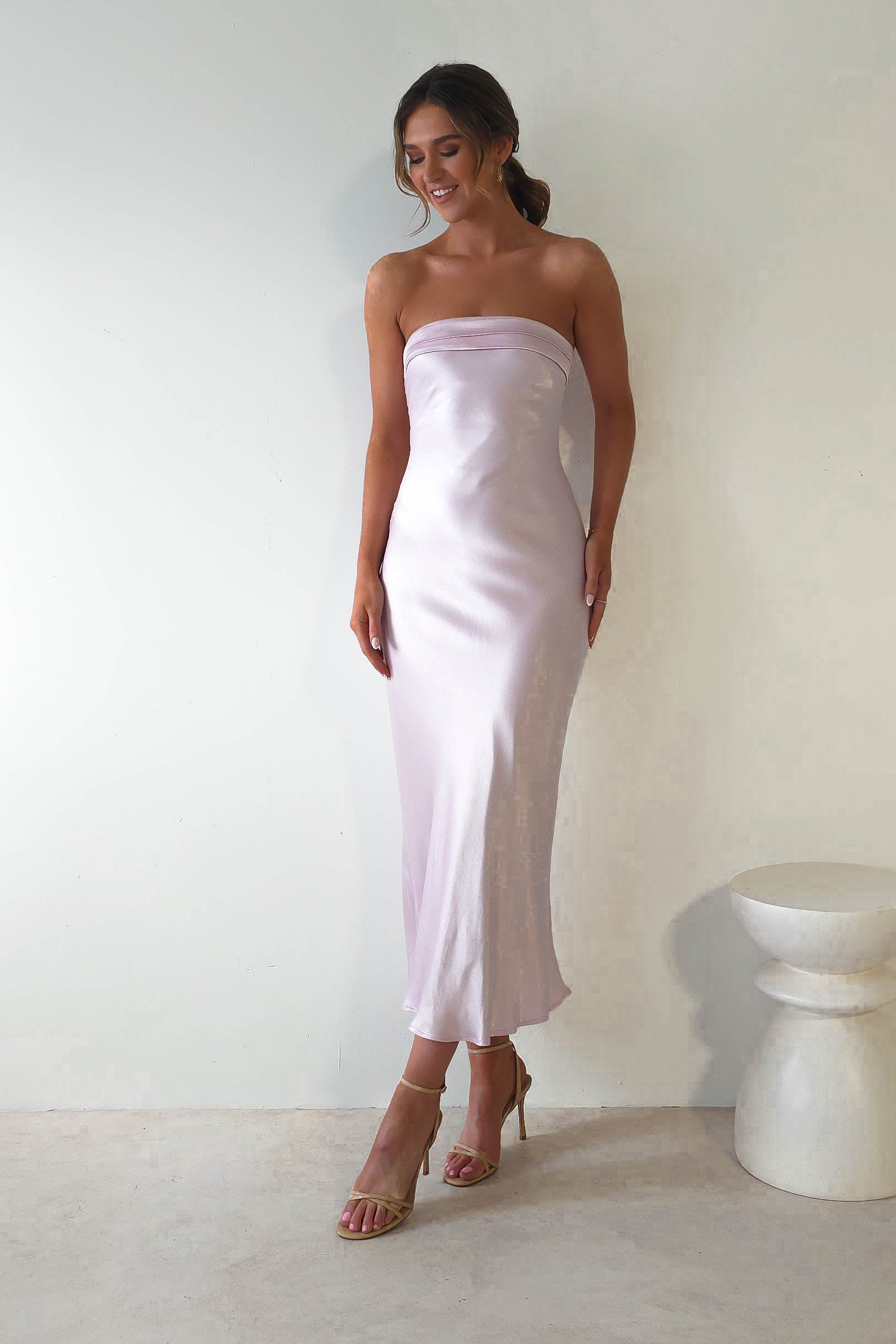 A woman in a strapless midi dress, made of silky super soft fabric, poses elegantly against a white wall. She is wearing strappy high heels and has her hair styled up. A small round white table is partially visible beside her, making for the perfect look for wedding guests.