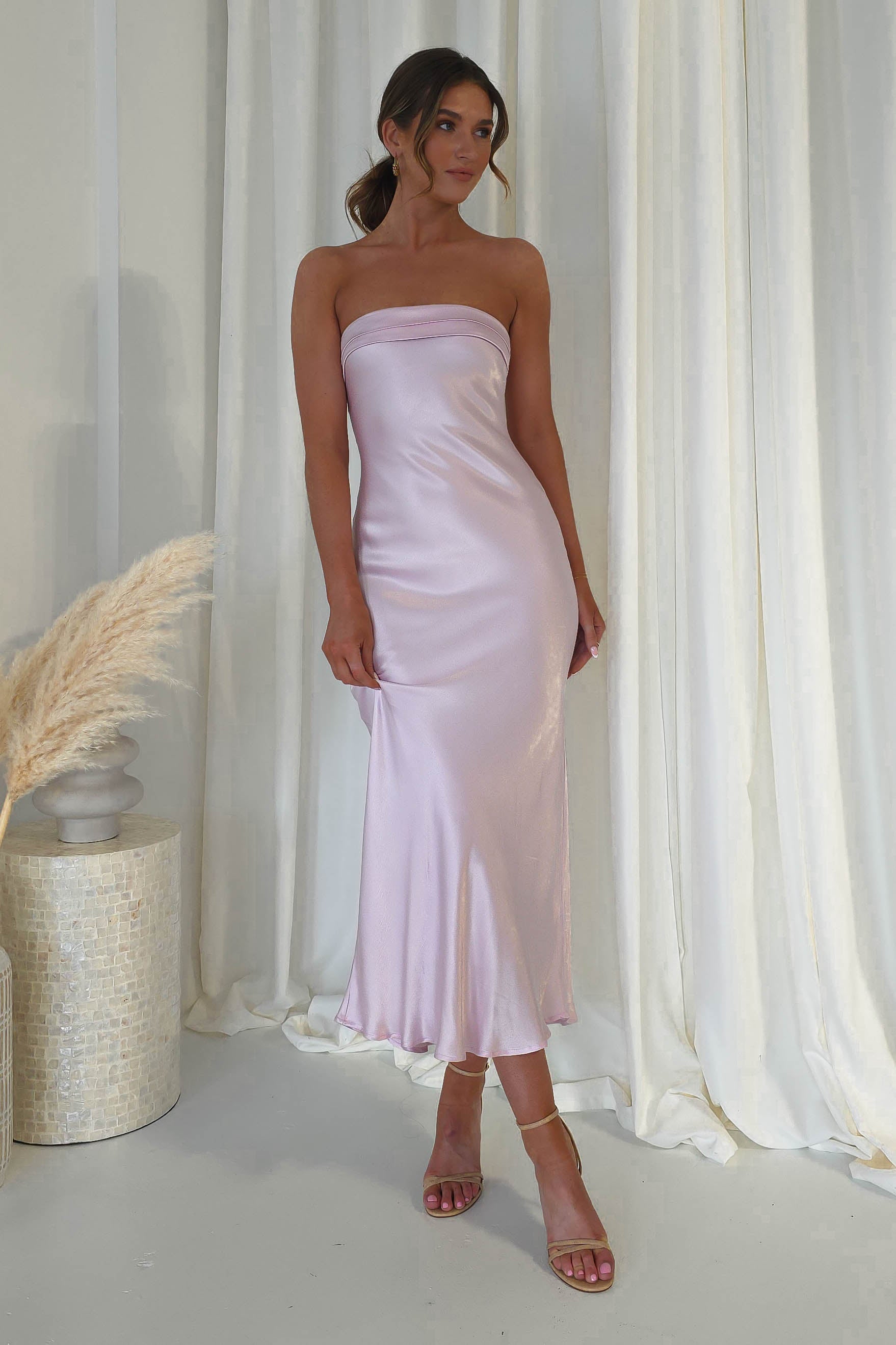 A wedding guest stands in a shiny, strapless midi dress against white curtains. Theyre wearing strappy high-heeled sandals and stand beside a tall vase with dried pampas grass.