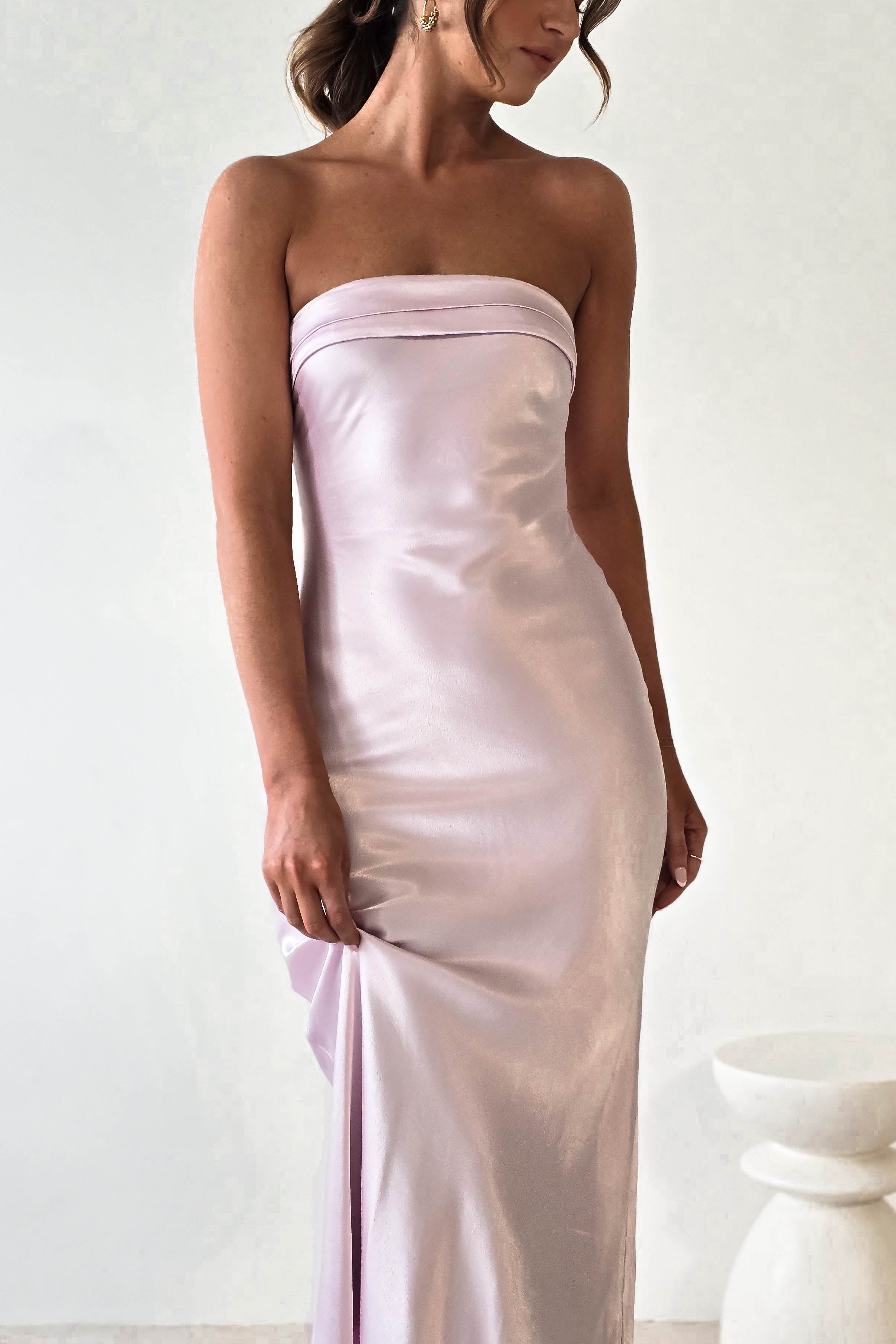 A woman wearing a strapless, shiny lavender midi dress stands in front of a plain white background. The gown, perfect for wedding guests, features a satin-like, super soft fabric. She is gently holding the material at her side, and her hair is styled in loose curls.