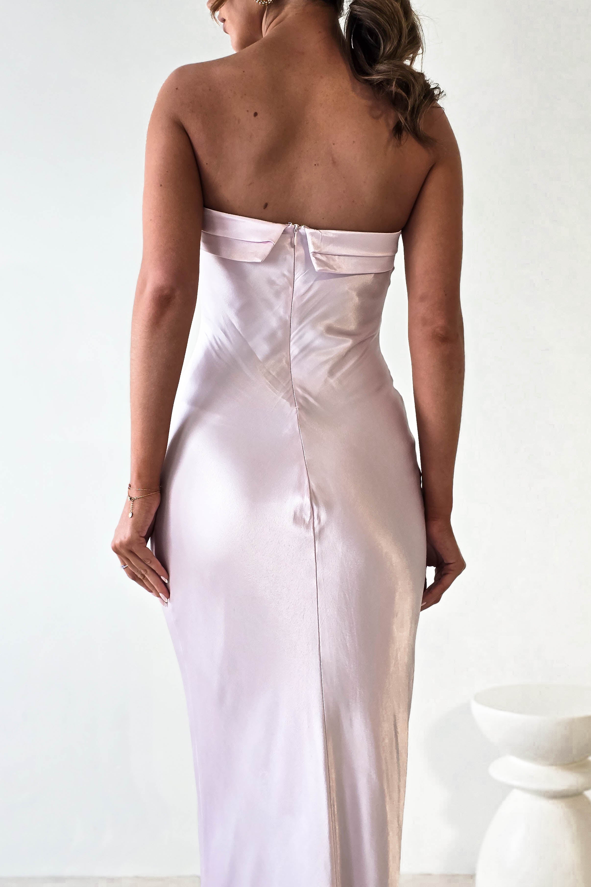 A person with long hair is wearing a strapless midi dress in light pink satin, crafted from super soft fabric. Standing with their back to the camera against a plain white background, they exude an elegance perfect for wedding guests.
