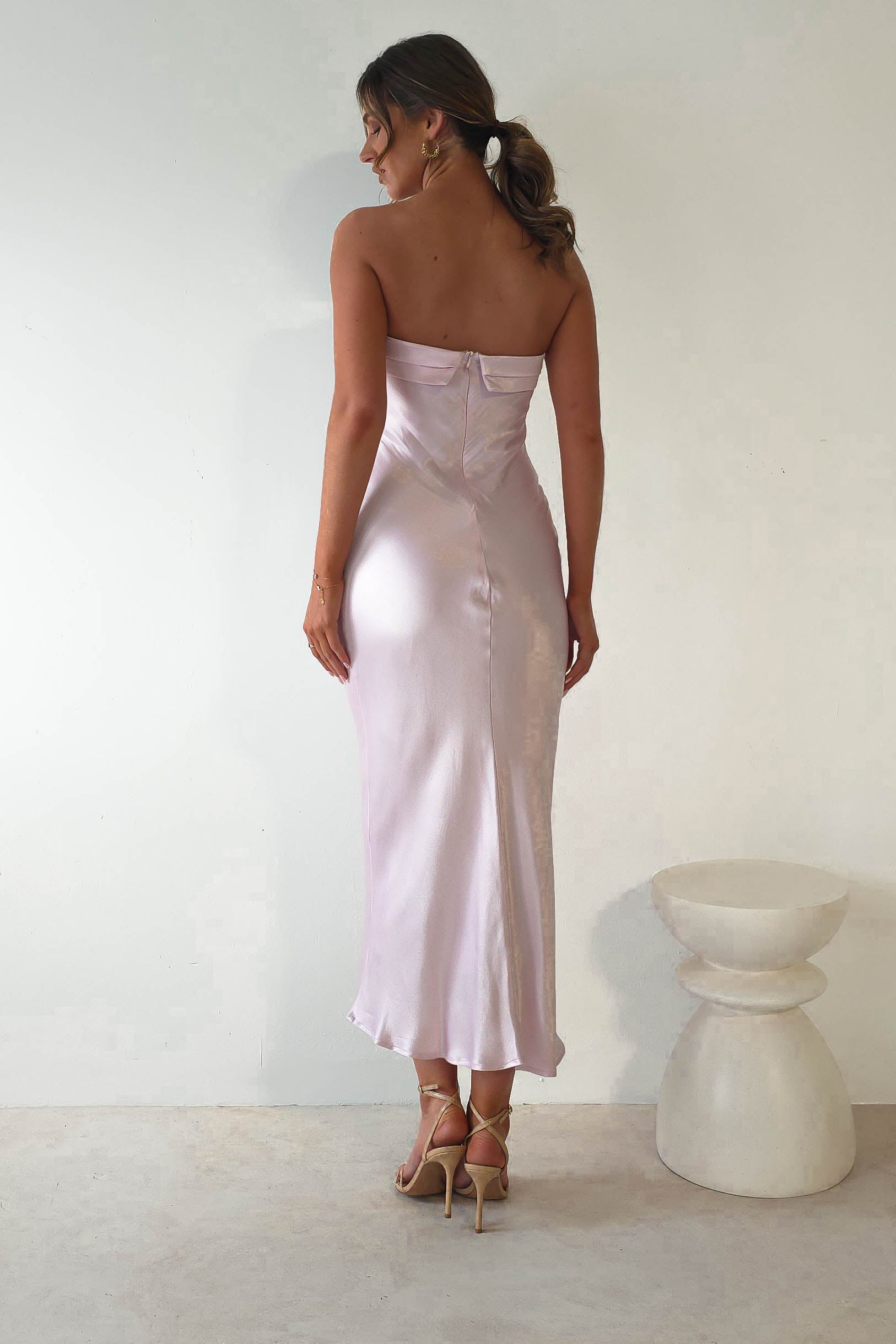 A woman wearing a strapless midi dress in light pink satin and strappy high heels stands with her back to the camera. Her hair is styled in a low ponytail, perfect for wedding guests. A small white pedestal is visible to her right.