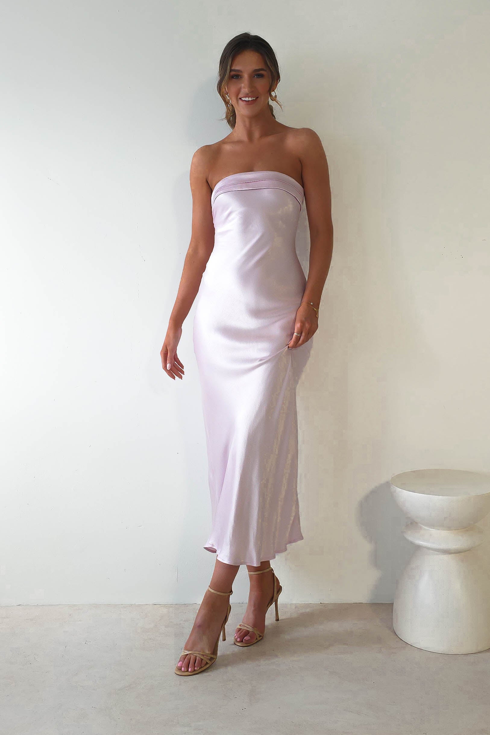 A woman stands against a light wall wearing a strapless midi dress in silky, light lavender fabric. Her hair is up, and shes adorned with dangling earrings and heels. A small, white, rounded stool is seen nearby, offering the perfect look for stylish wedding guests.