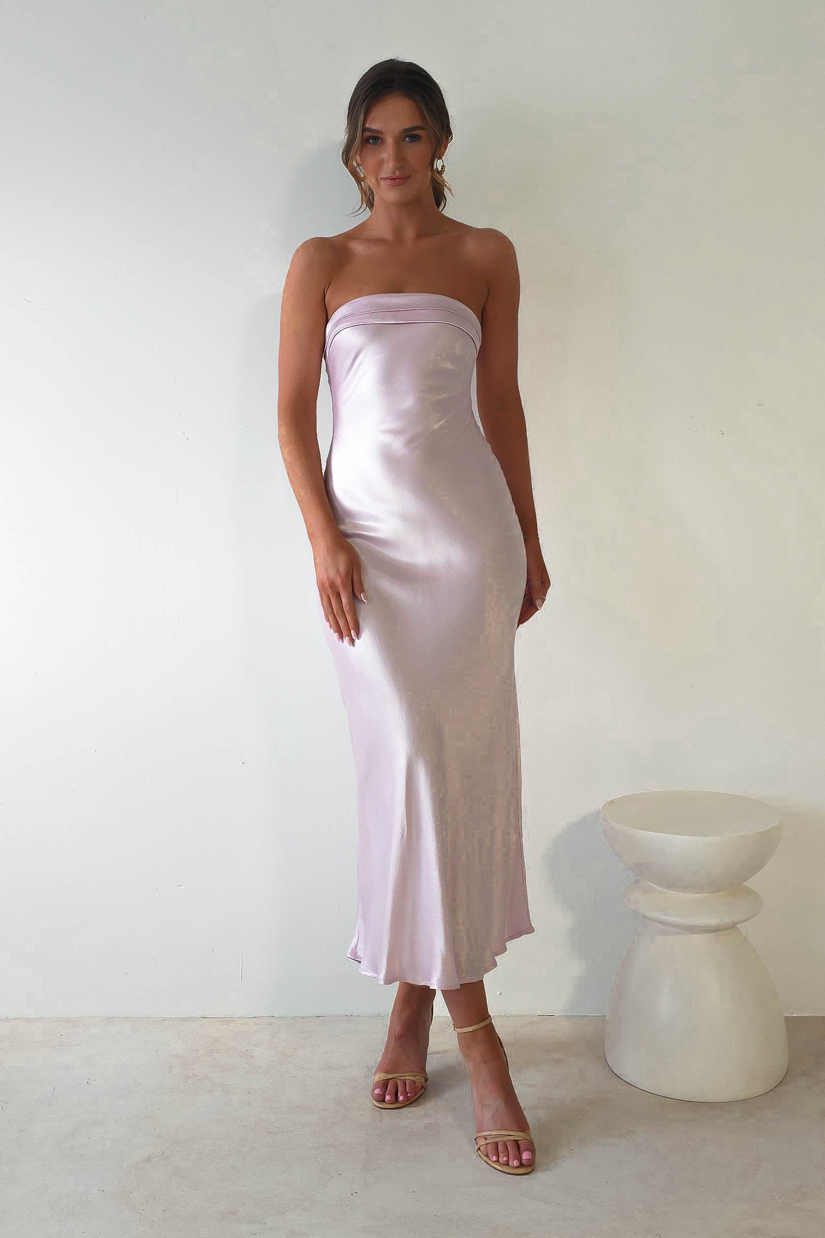 A person stands against a plain background wearing a silky, strapless midi dress in light lavender. They pair it elegantly with strappy high-heeled sandals. A round white pedestal is positioned to their right, perfect for attracting admiration from wedding guests.