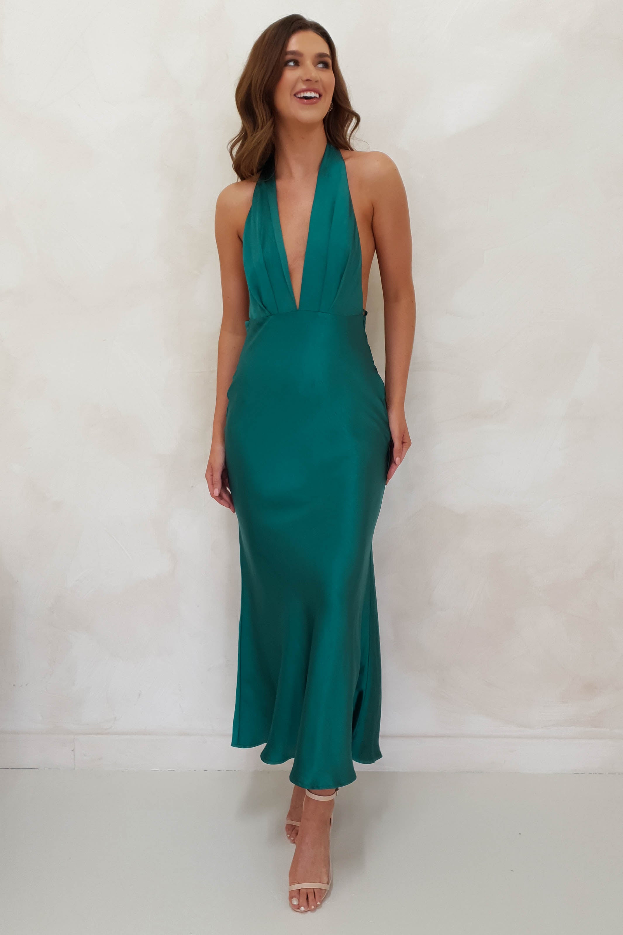 A woman in a teal, halter-neck satin dress smiles against a light background. Perfect for a wedding guest, the floor-length gown with a deep V-neckline elegantly complements her wavy brown hair and nude high-heeled sandals, all in soft satin material.
