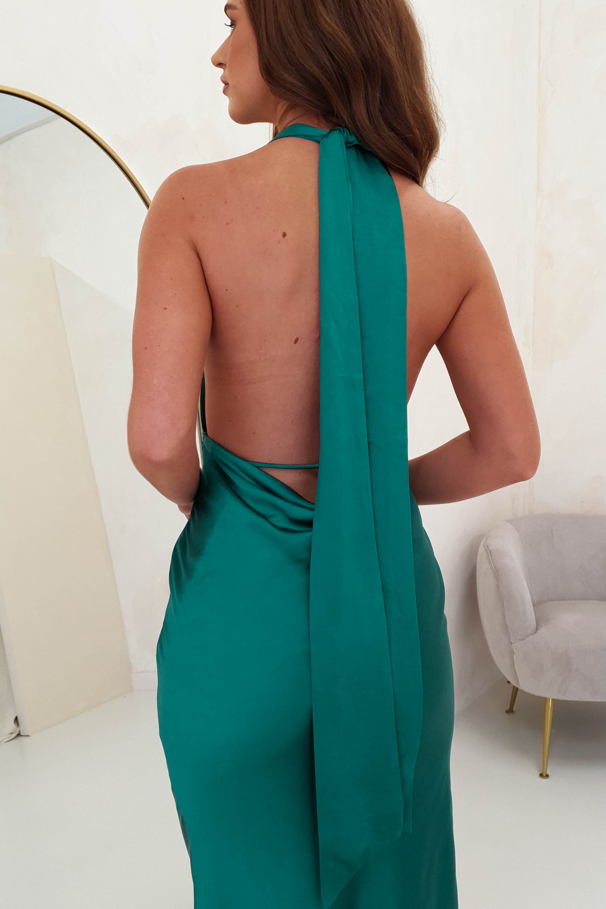 A woman with long brown hair is facing away, wearing a teal backless halter dress crafted from soft satin material. She stands near a light-colored chair in a minimalist room with light walls and a gold-framed mirror, the glamorous neck-tie of her dress elegantly draped down her back.