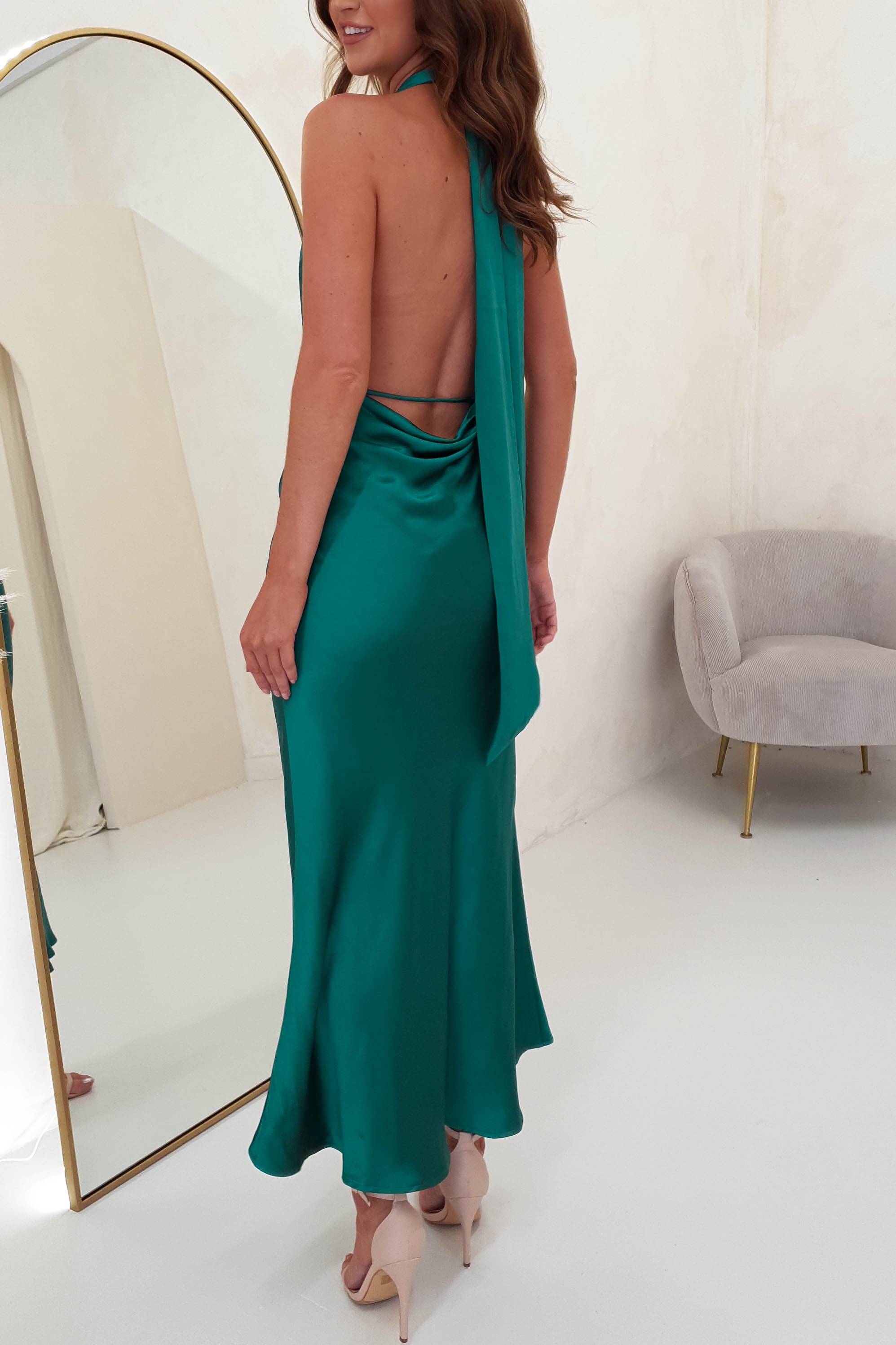 A woman, possibly a chic wedding guest, gazes at her reflection in a full-length mirror. She wears a backless teal satin dress made from soft satin material, with long brown hair cascading over her shoulders. Nearby is a light gray chair with gold legs on the white floor.