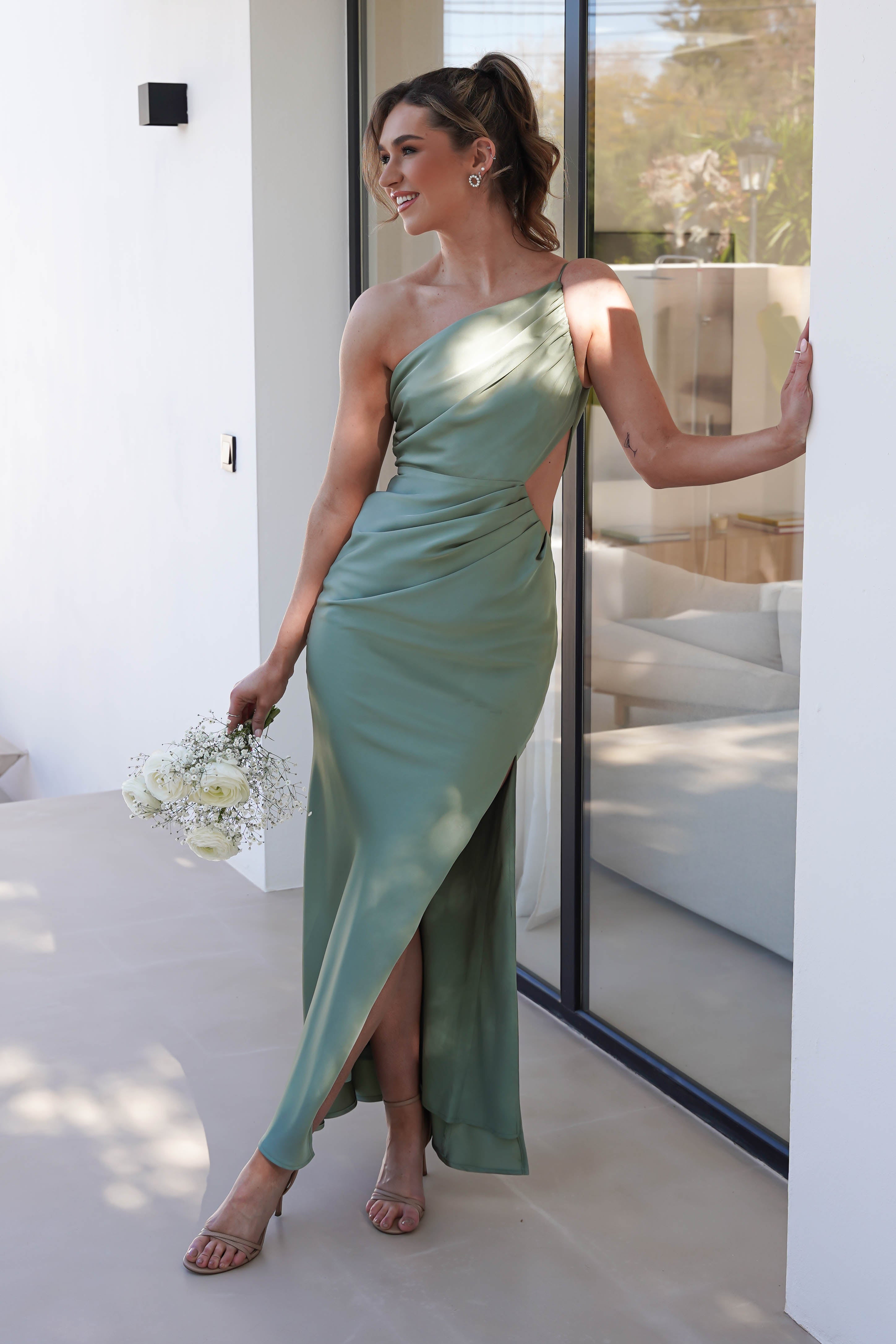 A woman in a green satin maxi dress with a side slit stands by a glass door, holding a bouquet of white flowers. The one-shoulder dress elegantly complements her high heels, as she smiles and looks to the side. Sunlight casts soft shadows on the floor, enhancing the wedding guest dress ambiance.