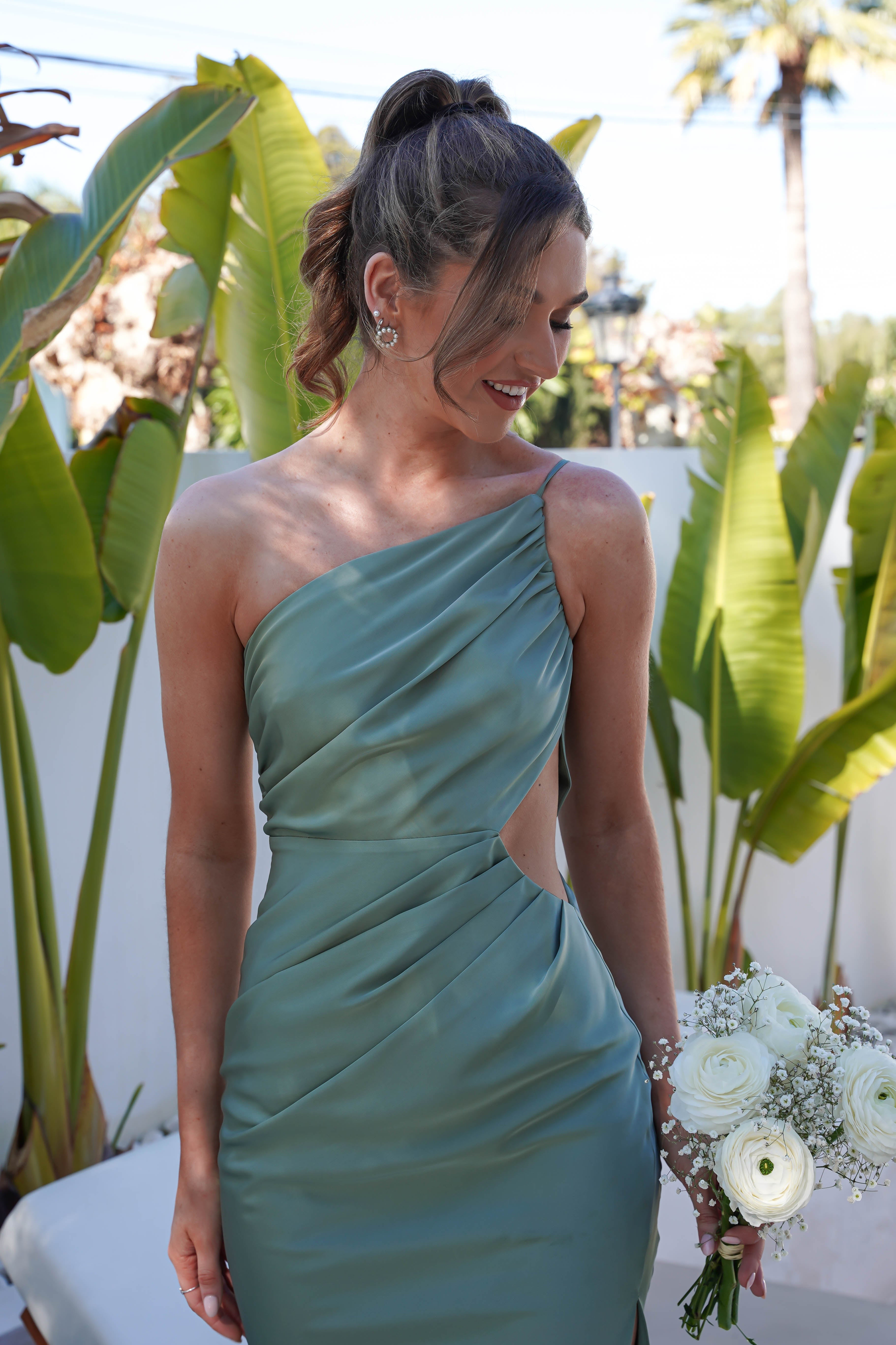 A woman in a green satin maxi dress holds a bouquet of white flowers, smiling as she gazes to the side. In the background, large green leaves contrast against a white wall.