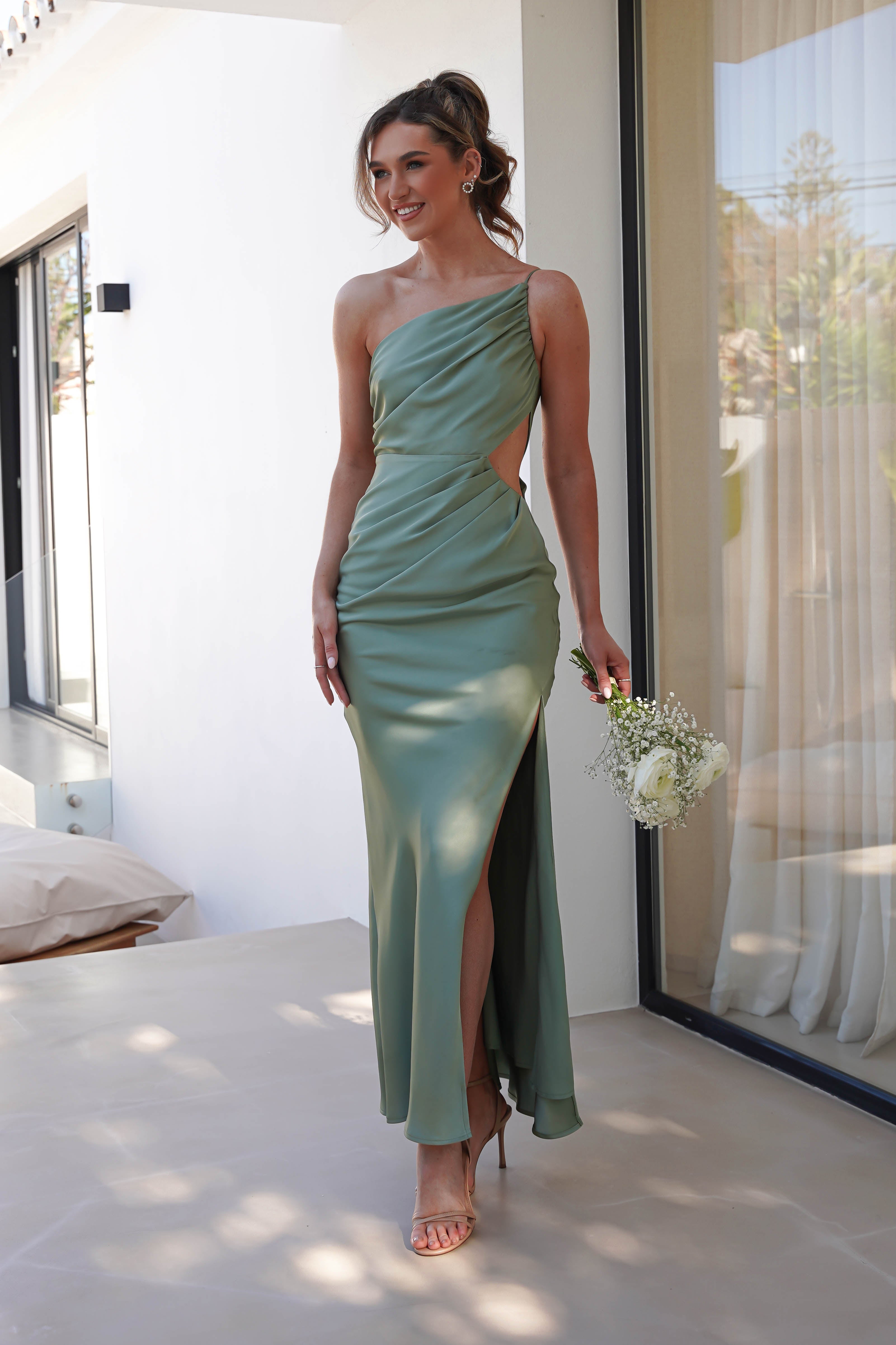 A woman in a green satin maxi dress gracefully stands on a patio, holding a bouquet of white flowers. The one-shoulder dress features side cutouts and a thigh-high slit. She complements her wedding guest look with beige high heels and an elegant updo.