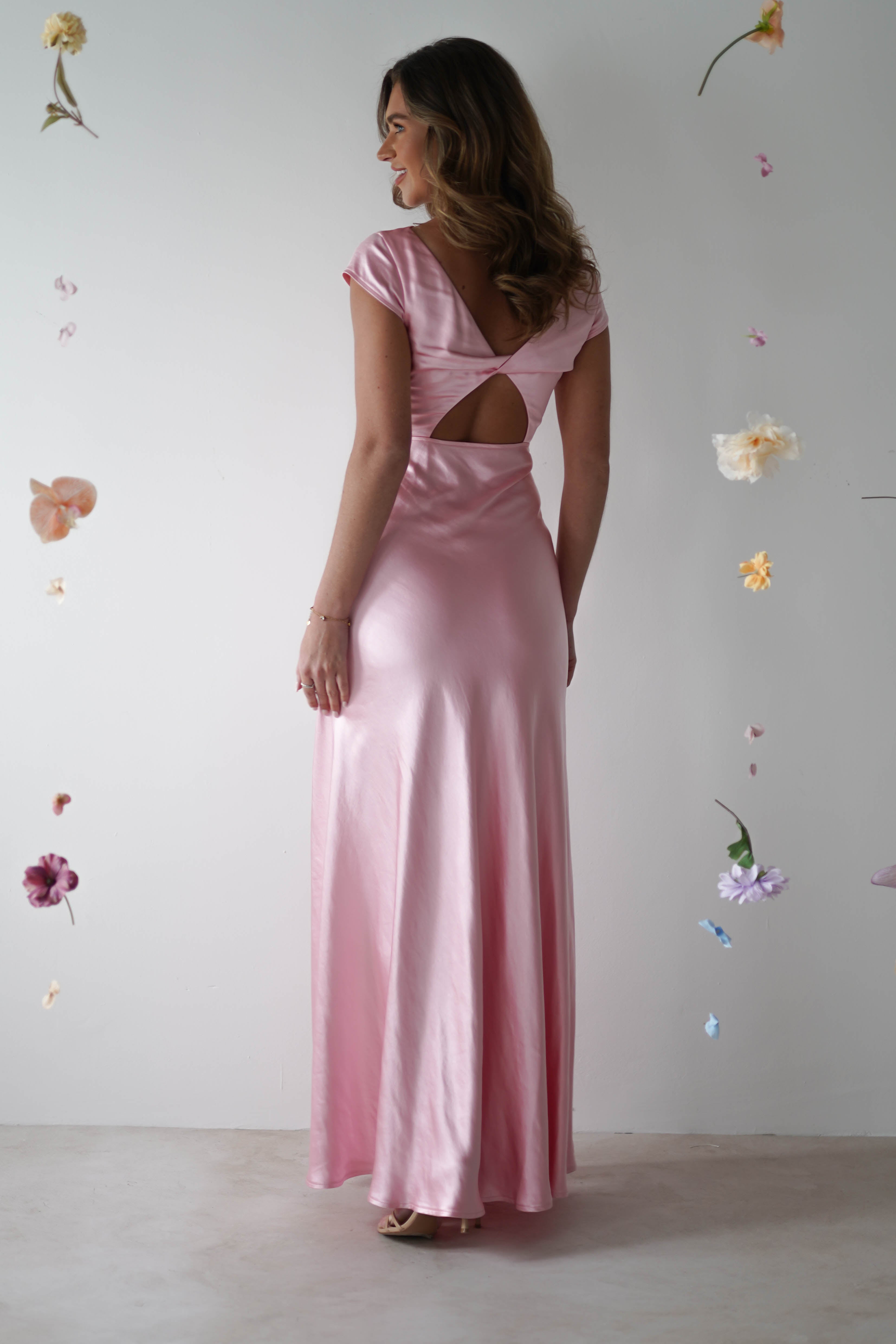 A woman in a long, pink maxi slip dress with a cutout back stands facing away, surrounded by floating pastel flowers against a plain white background—perfect inspiration for chic bridesmaids looks.