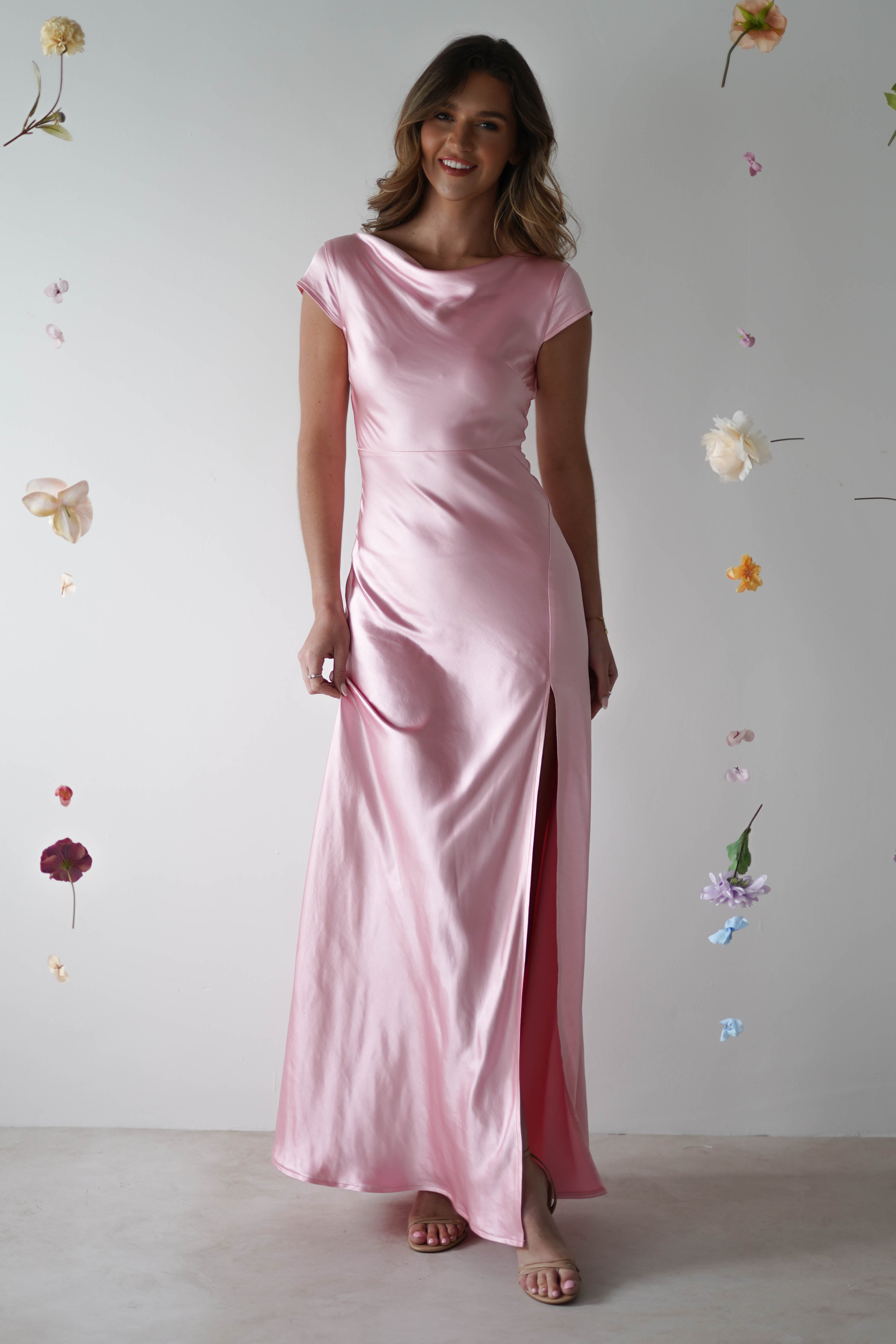 A woman wearing a shiny, light pink satin maxi slip dress with short sleeves and a thigh-high slit stands against a plain background decorated with hanging flowers. She smiles softly, holding one side of her bridesmaid-style gown.