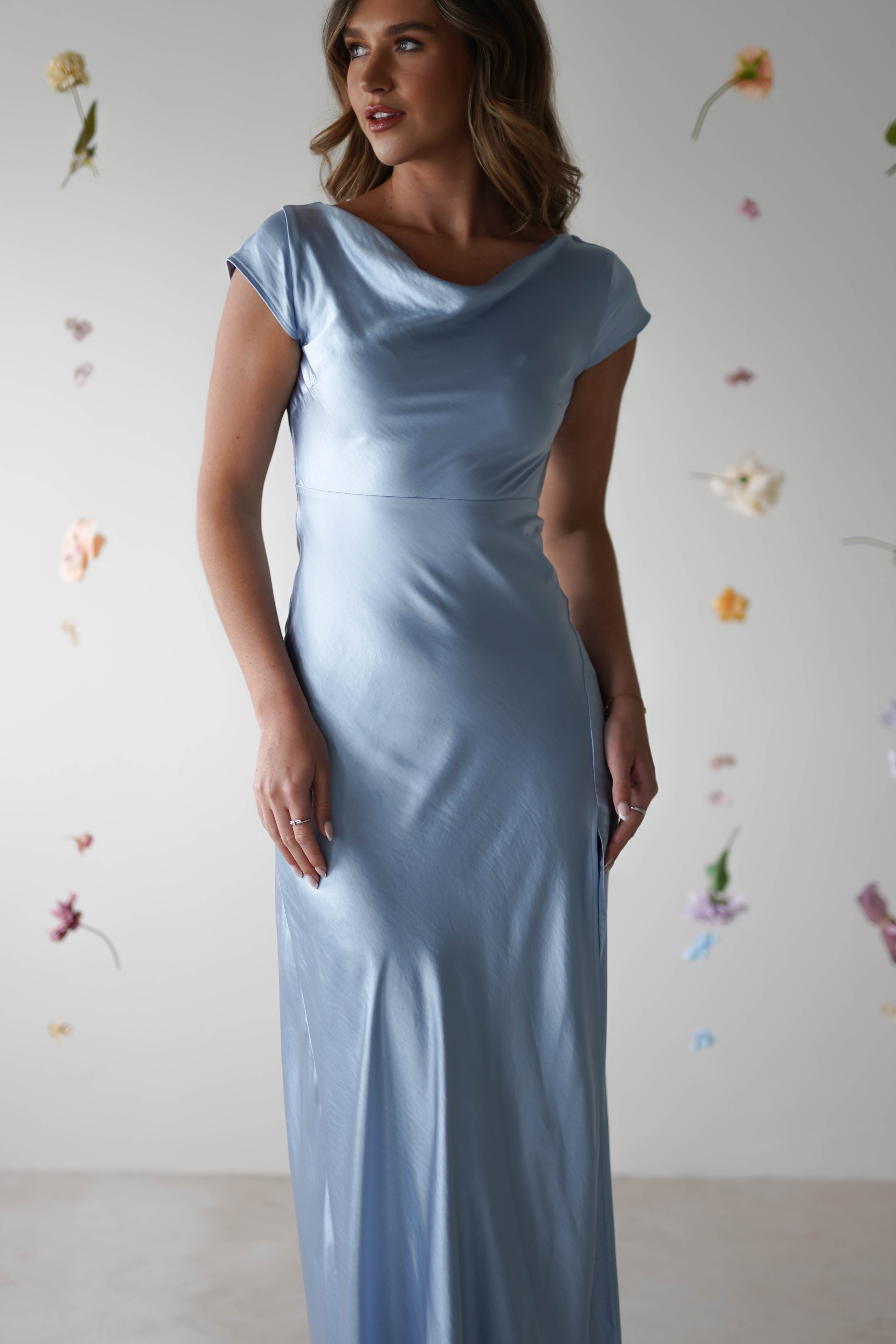 A woman wearing a silky light blue, figure slimming maxi slip dress with cap sleeves and a cowl neckline stands in front of a white background decorated with hanging, scattered flowers.