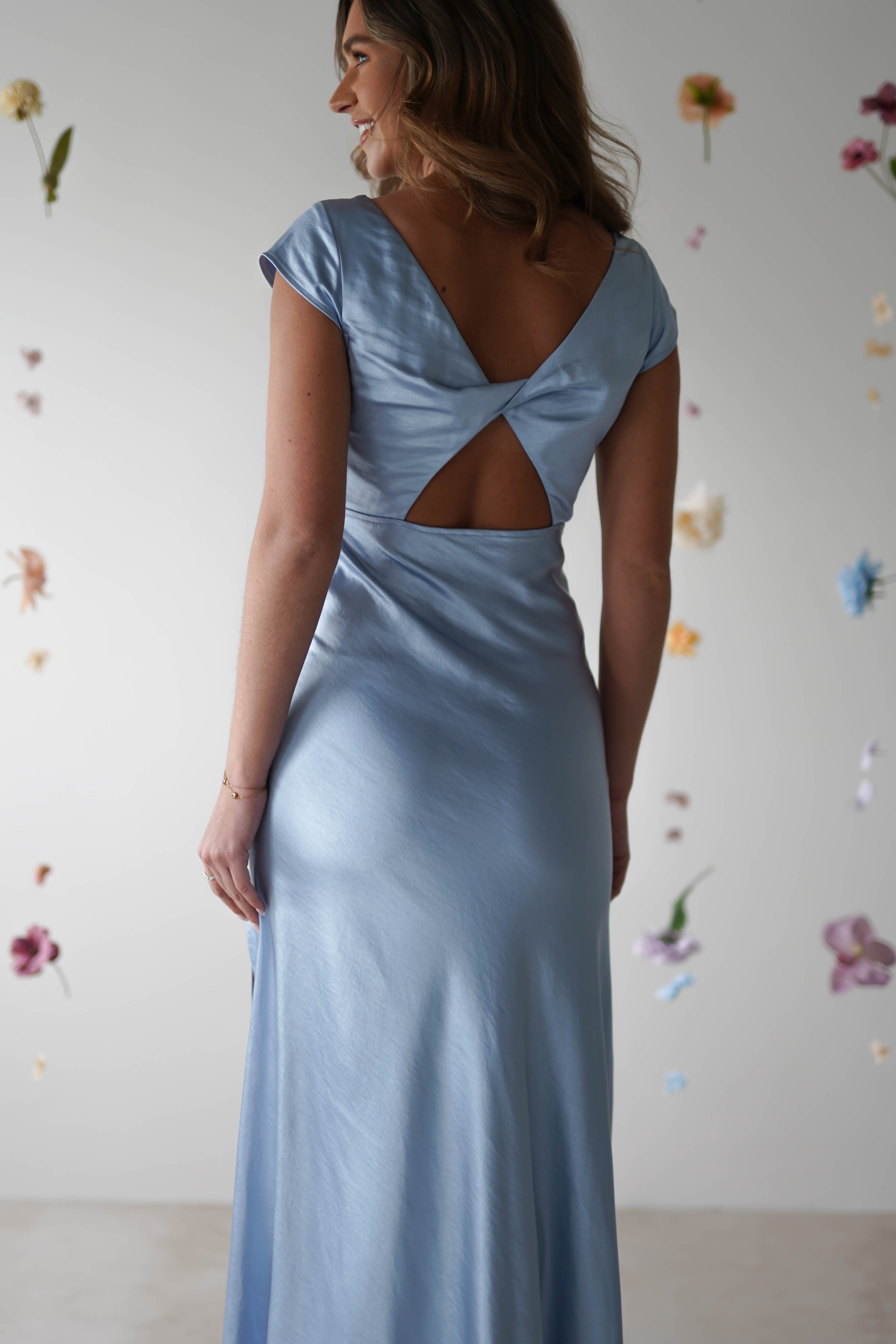 A woman in a light blue, figure slimming maxi slip dress with a V-shaped back and twisted back detail stands facing away, in front of a light-colored wall decorated with scattered, colorful artificial flowers.