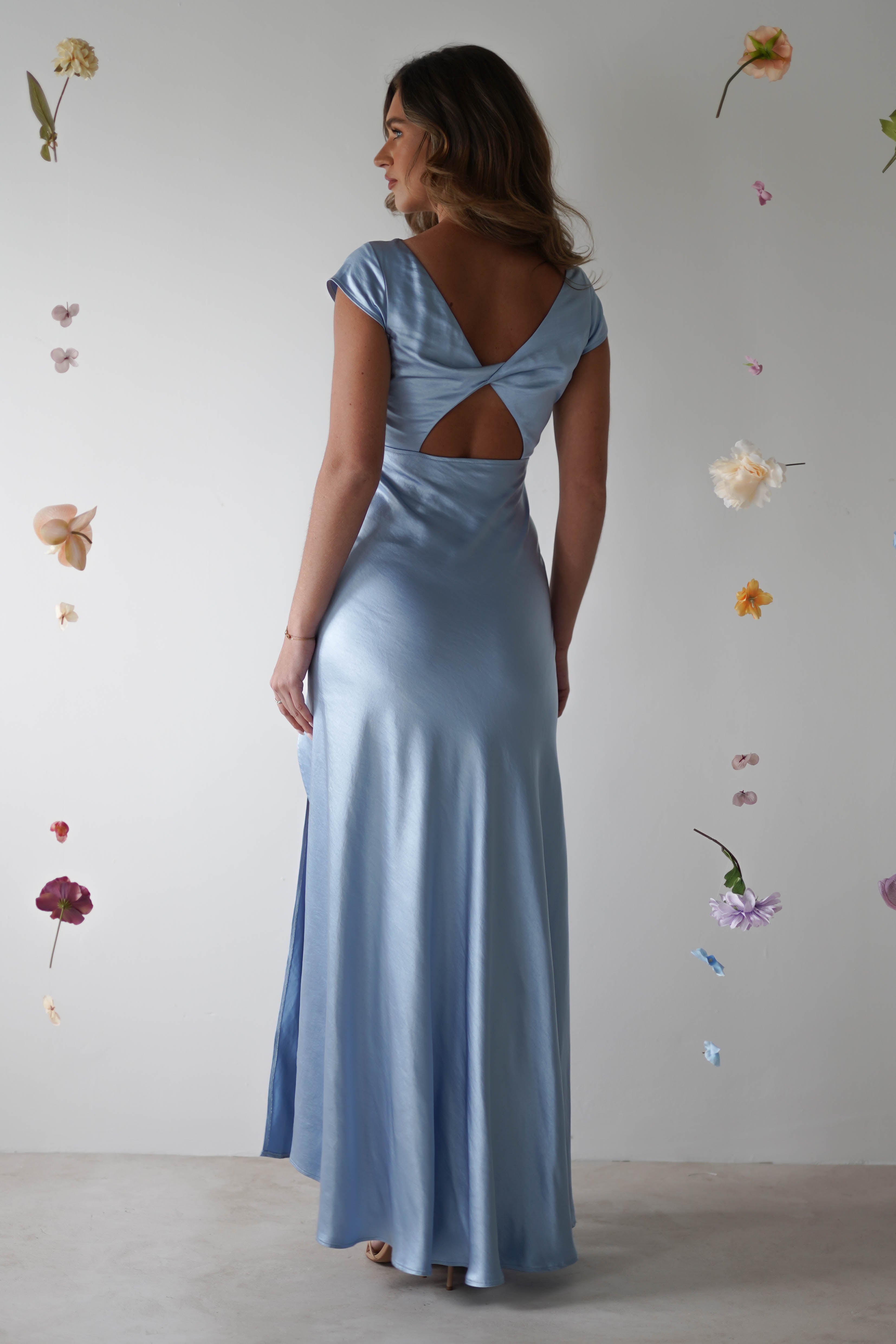 A woman in a light blue, figure slimming maxi slip dress with a cowl neckline and twisted back detail stands facing away, surrounded by floating, colorful flowers against a plain white background.