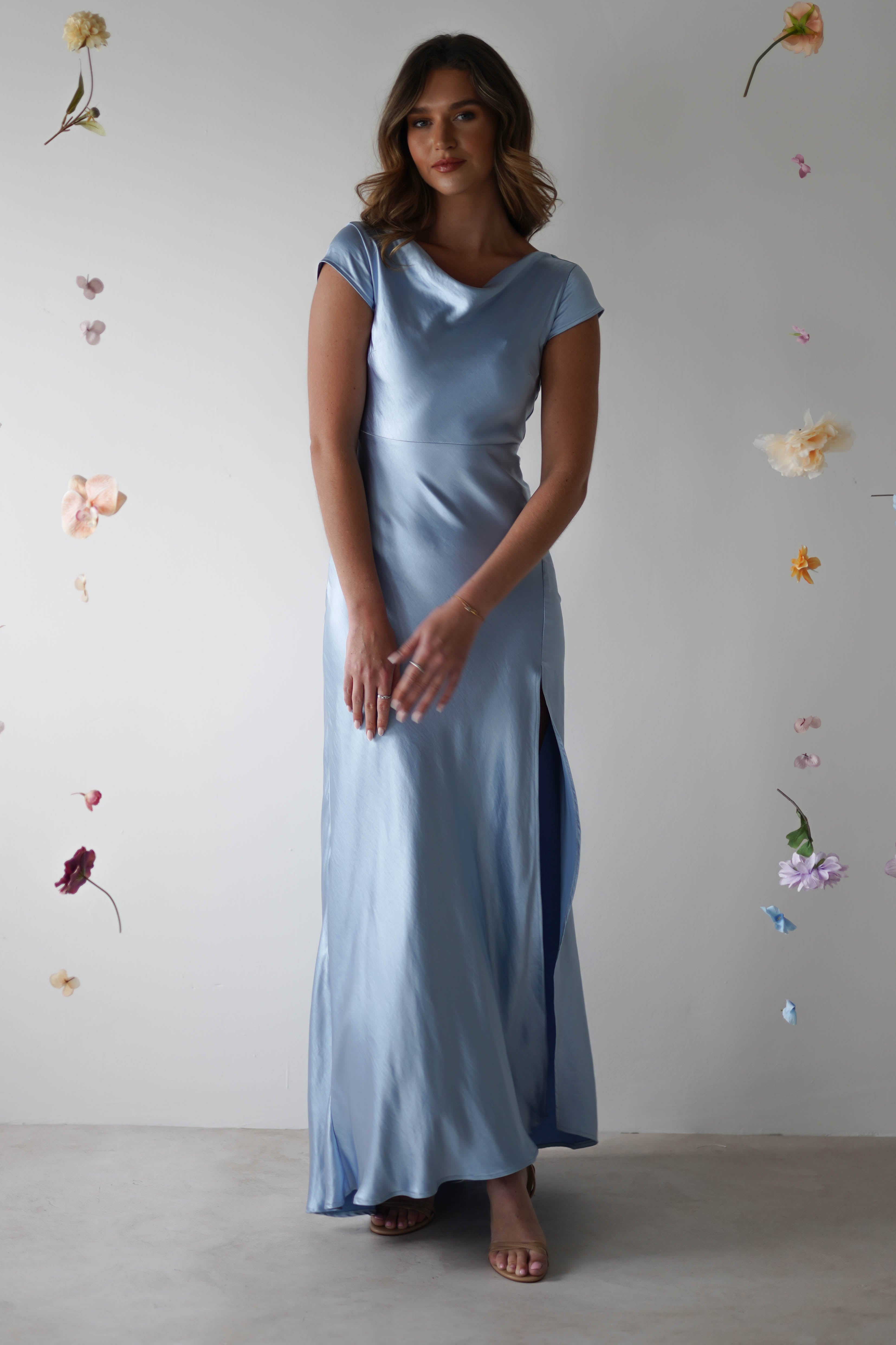 A woman in a light blue, figure slimming maxi slip dress with a cowl neckline stands against a white background adorned with small, colorful flowers. She looks at the camera, hands gently resting in front of her.