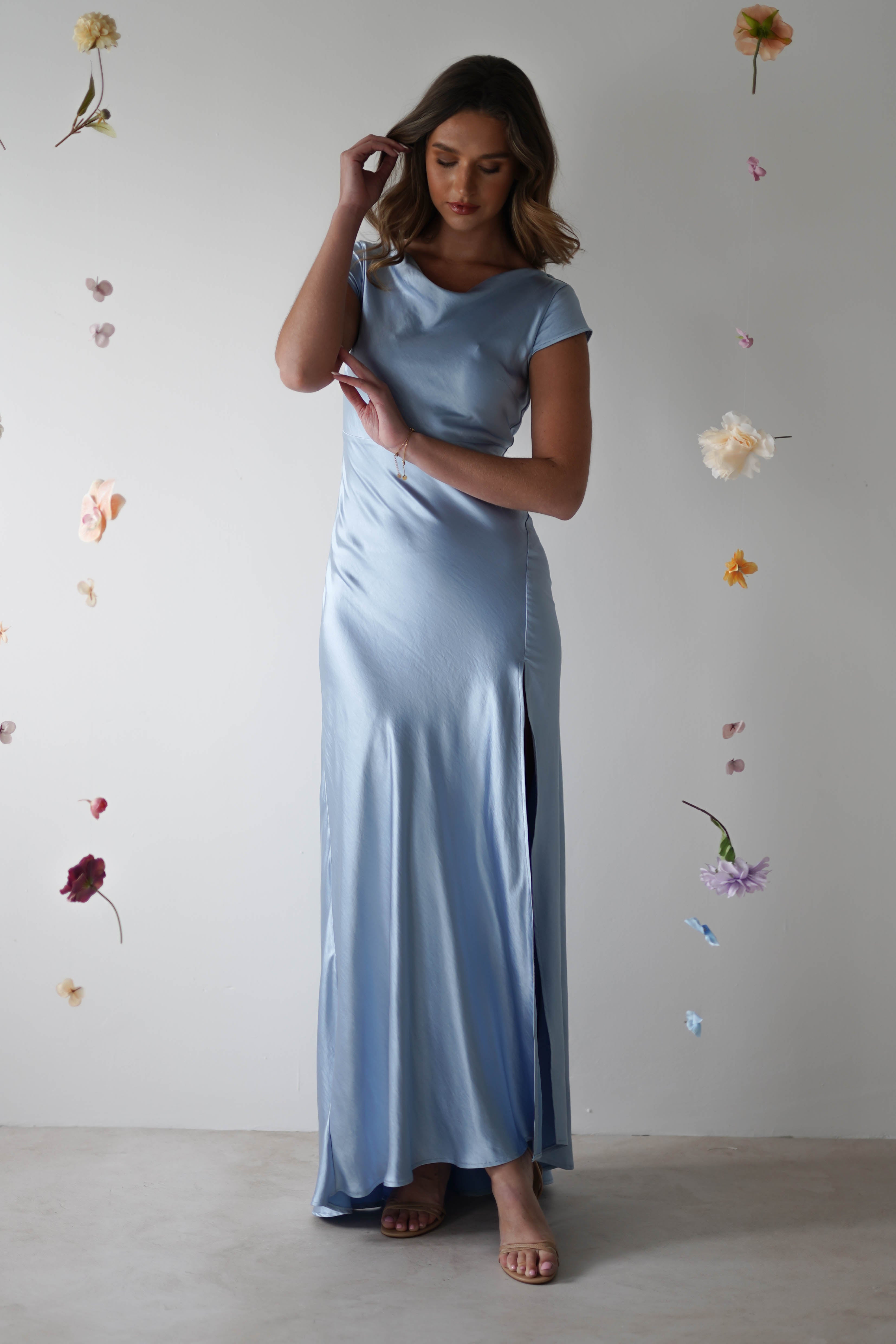 A woman in a light blue figure slimming maxi slip dress with a cowl neckline stands against a white background, surrounded by pastel flowers. One hand rests on her head, the other on her arm, as she gazes slightly downward.