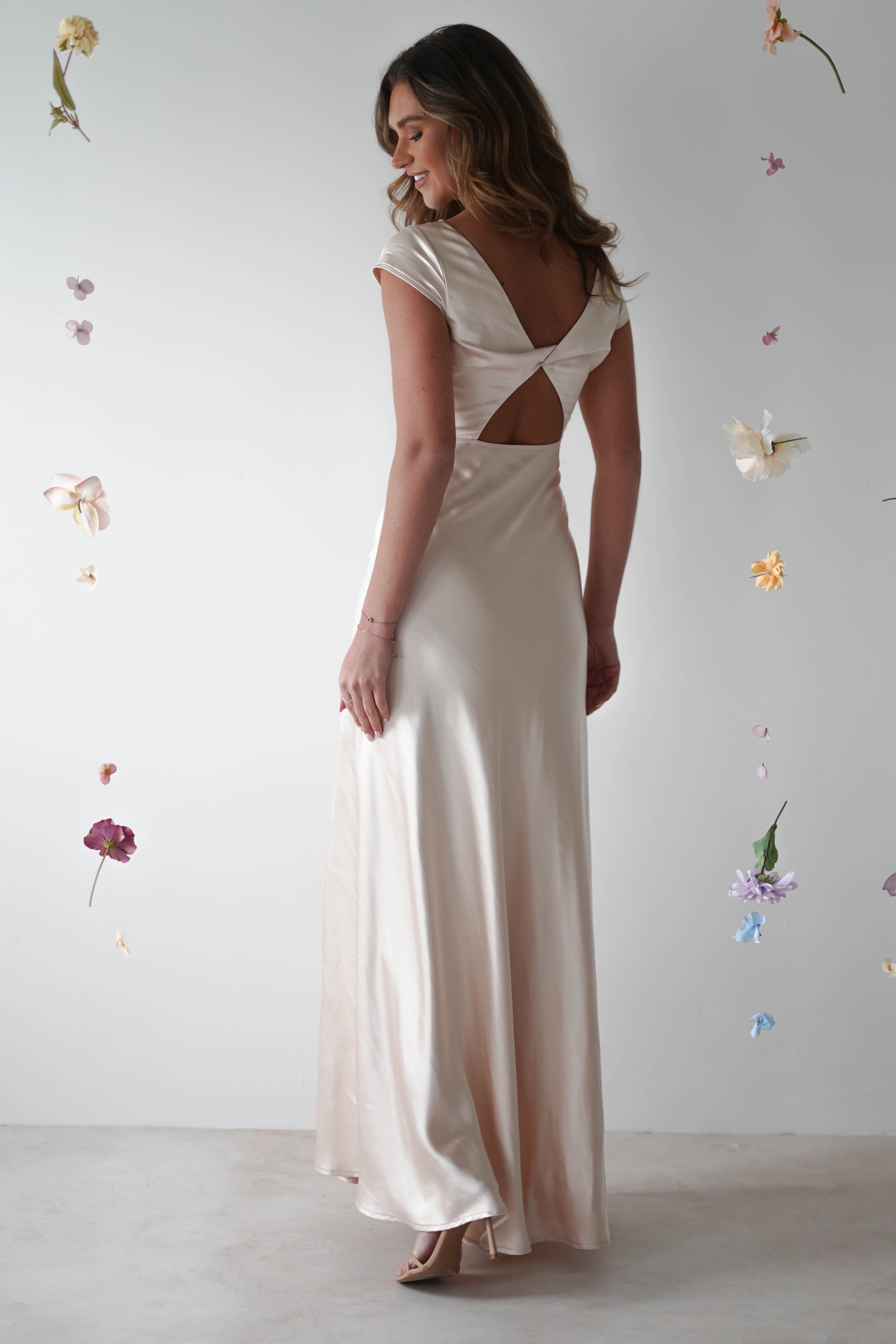 A woman in a long, elegant, cream-colored maxi slip dress with a cutout back stands facing away. Delicate flowers and petals appear to float around her against a plain, light background—perfect for special occasions or bridesmaids.