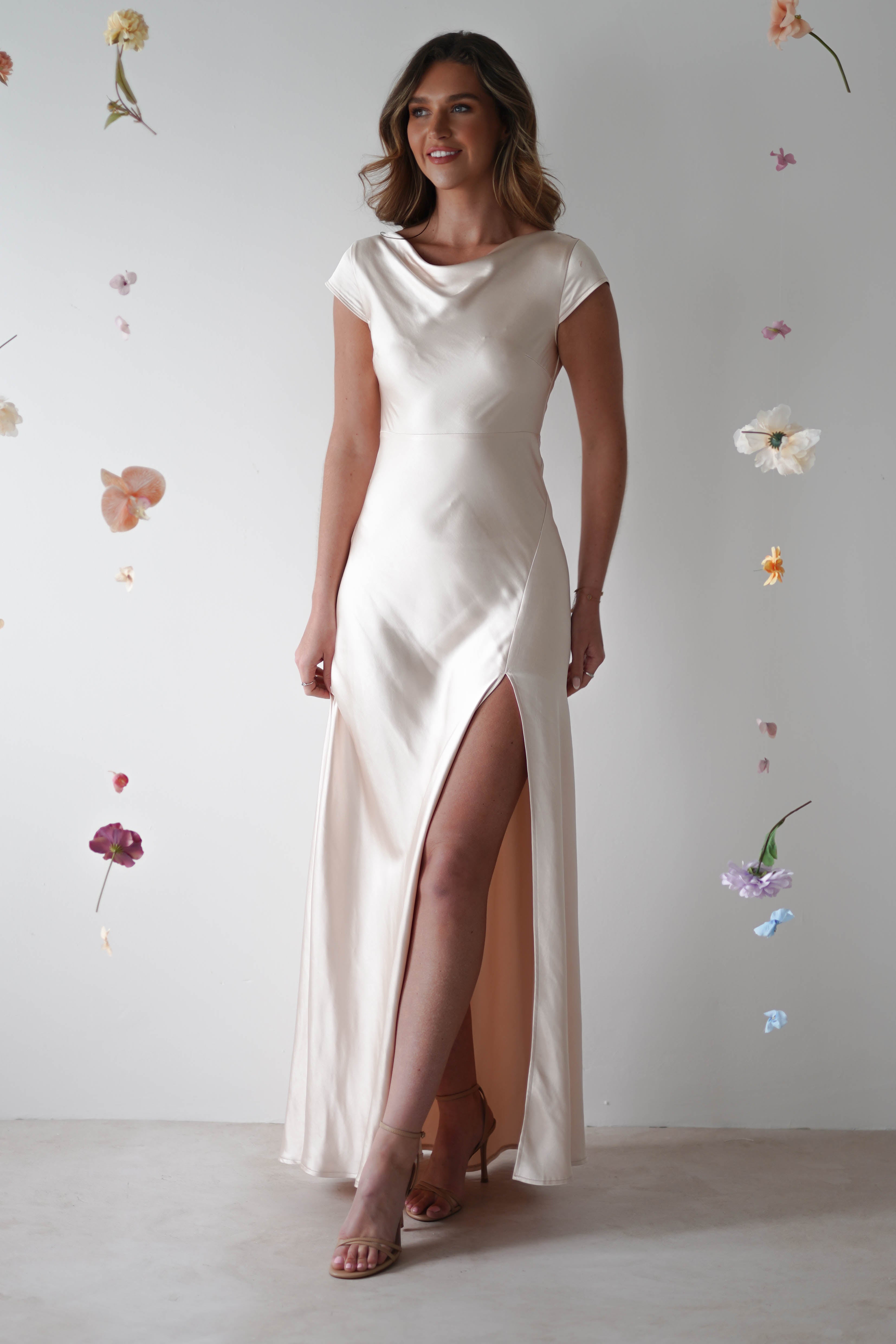 A woman in a silky ivory maxi slip dress with a thigh-high slit stands against a light backdrop with floating flowers. Perfect for special occasions or bridesmaids, she smiles slightly, her loose hair complementing her strappy heels.