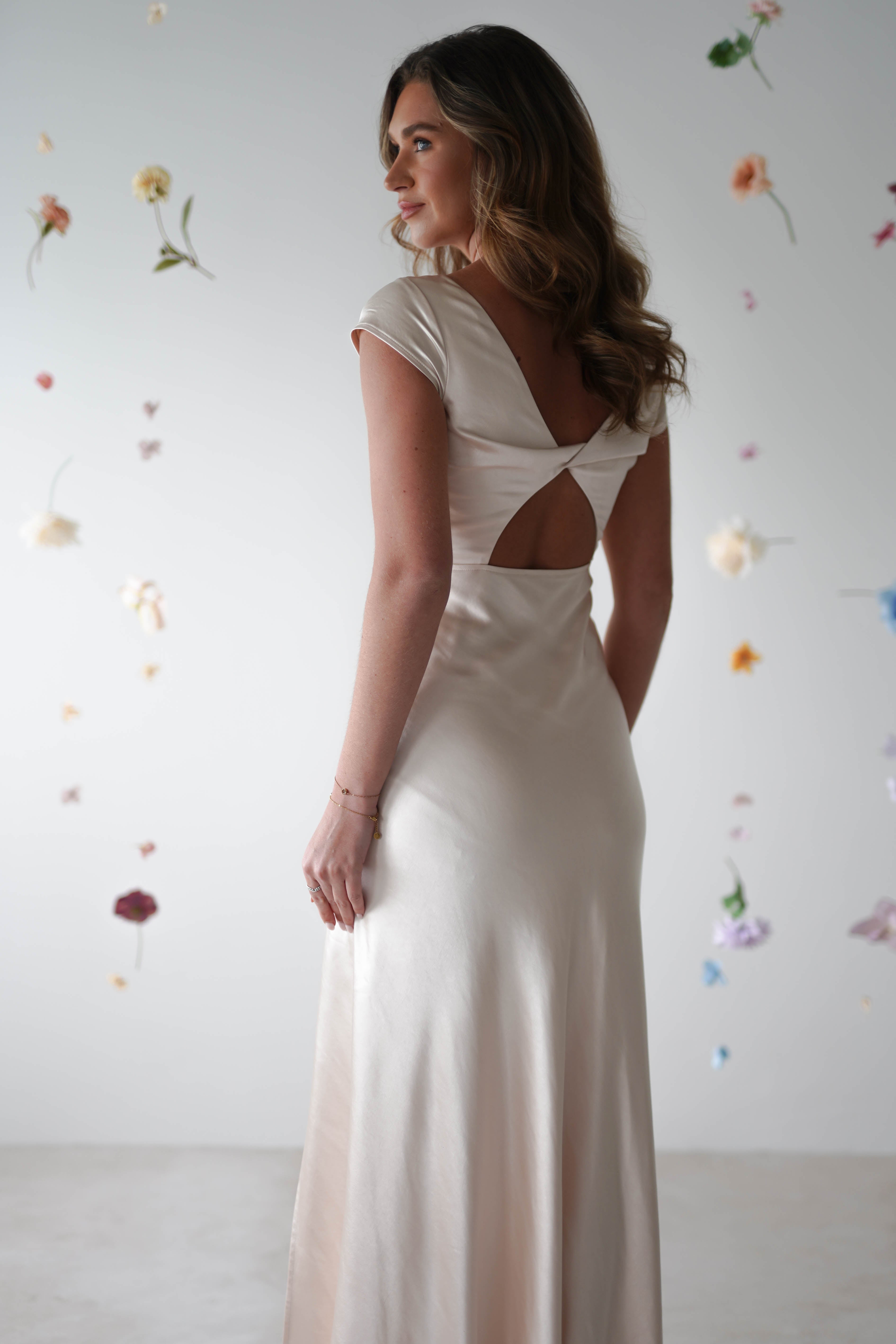 A woman in a white satin maxi slip dress with an open back stands facing away, surrounded by floating pastel flowers—a dreamy look perfect for bridesmaids or special occasions against a light background.
