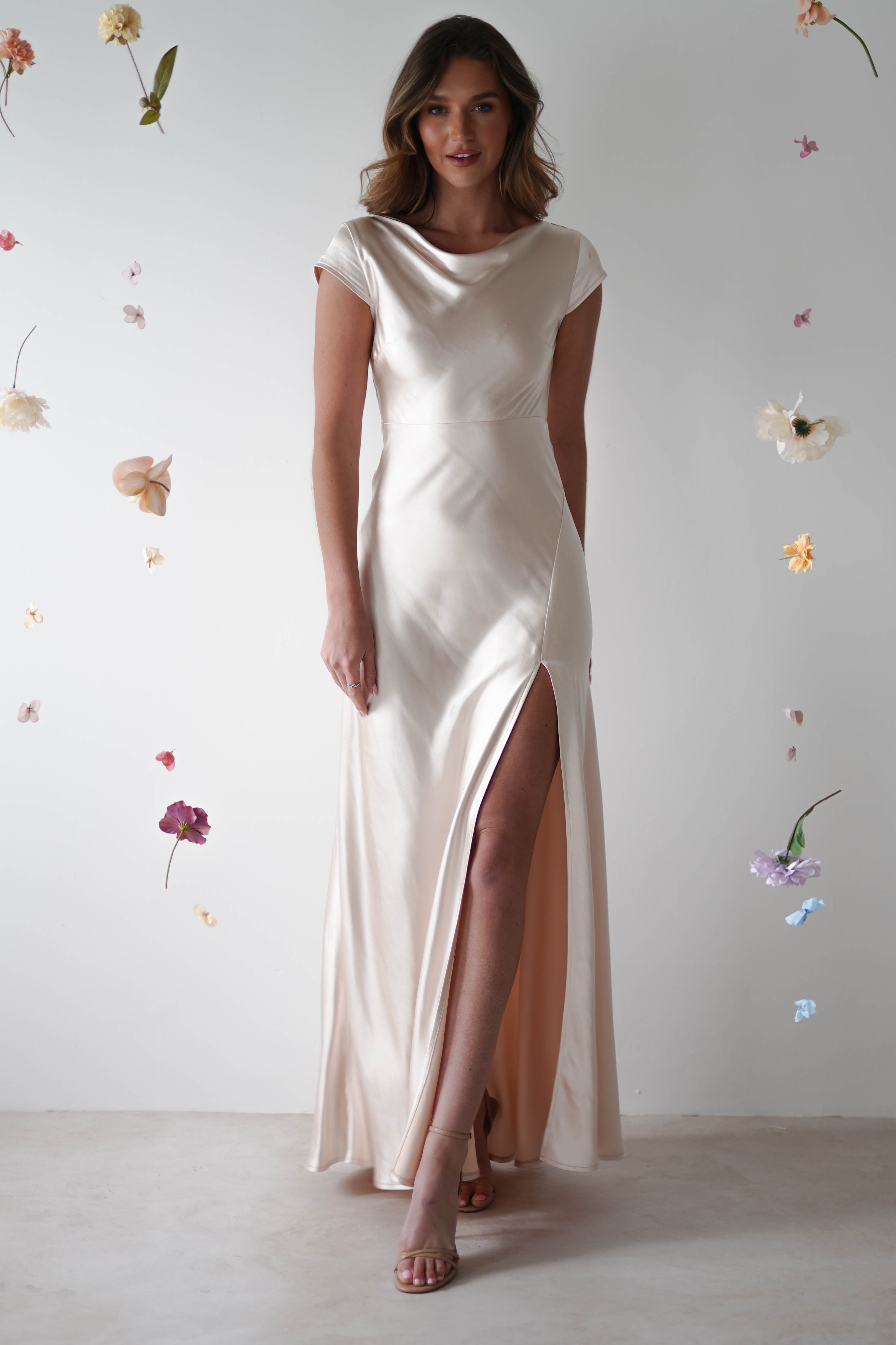 A woman in a silky, off-white maxi slip dress with a thigh-high slit stands against a white background, surrounded by floating pastel flowers—perfect for bridesmaids or special occasions.
