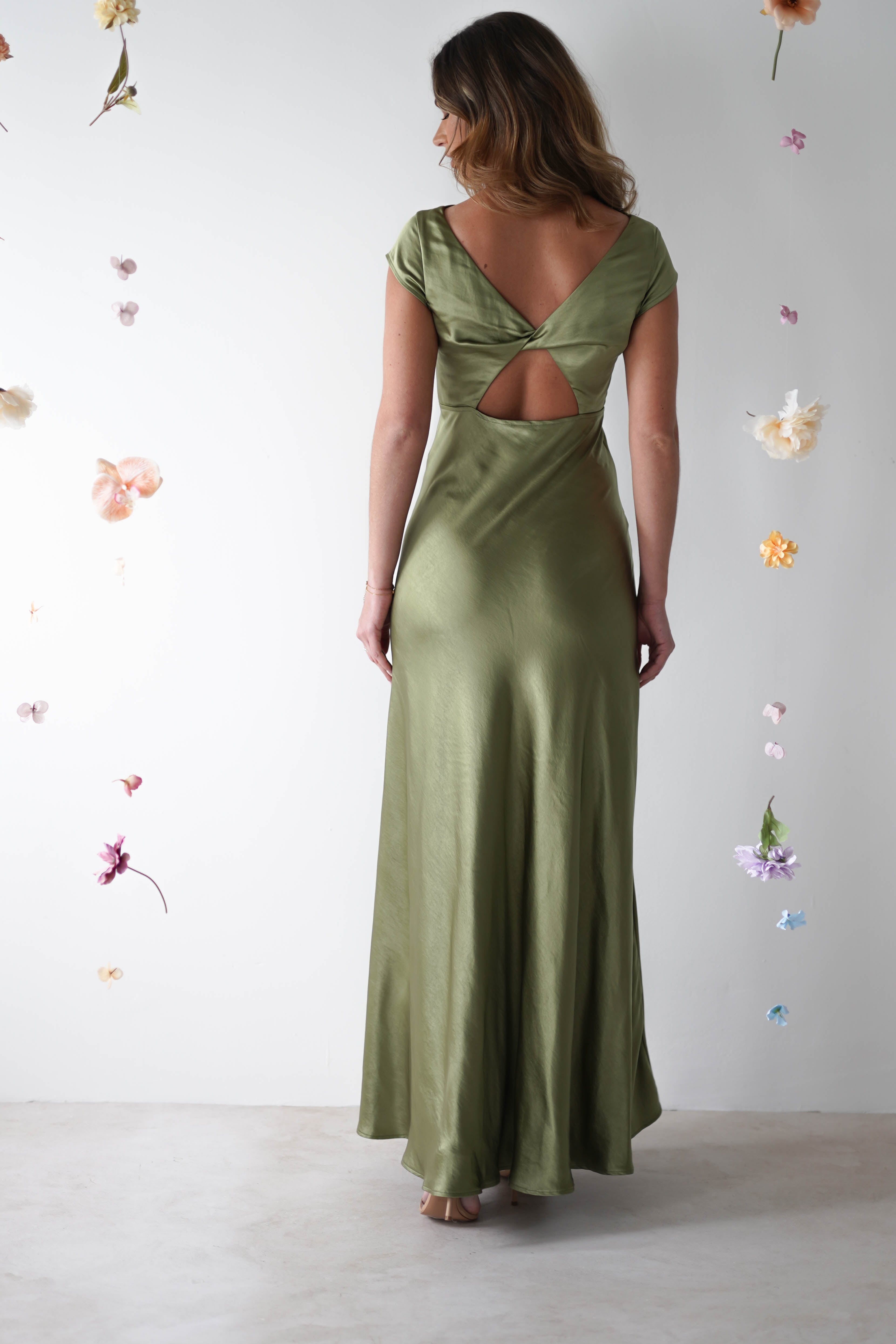 A woman in an elegant green maxi slip dress stands with her back turned, revealing a V-shaped open back and twisted back detail. She is surrounded by small, suspended colorful flowers on a light gray background.