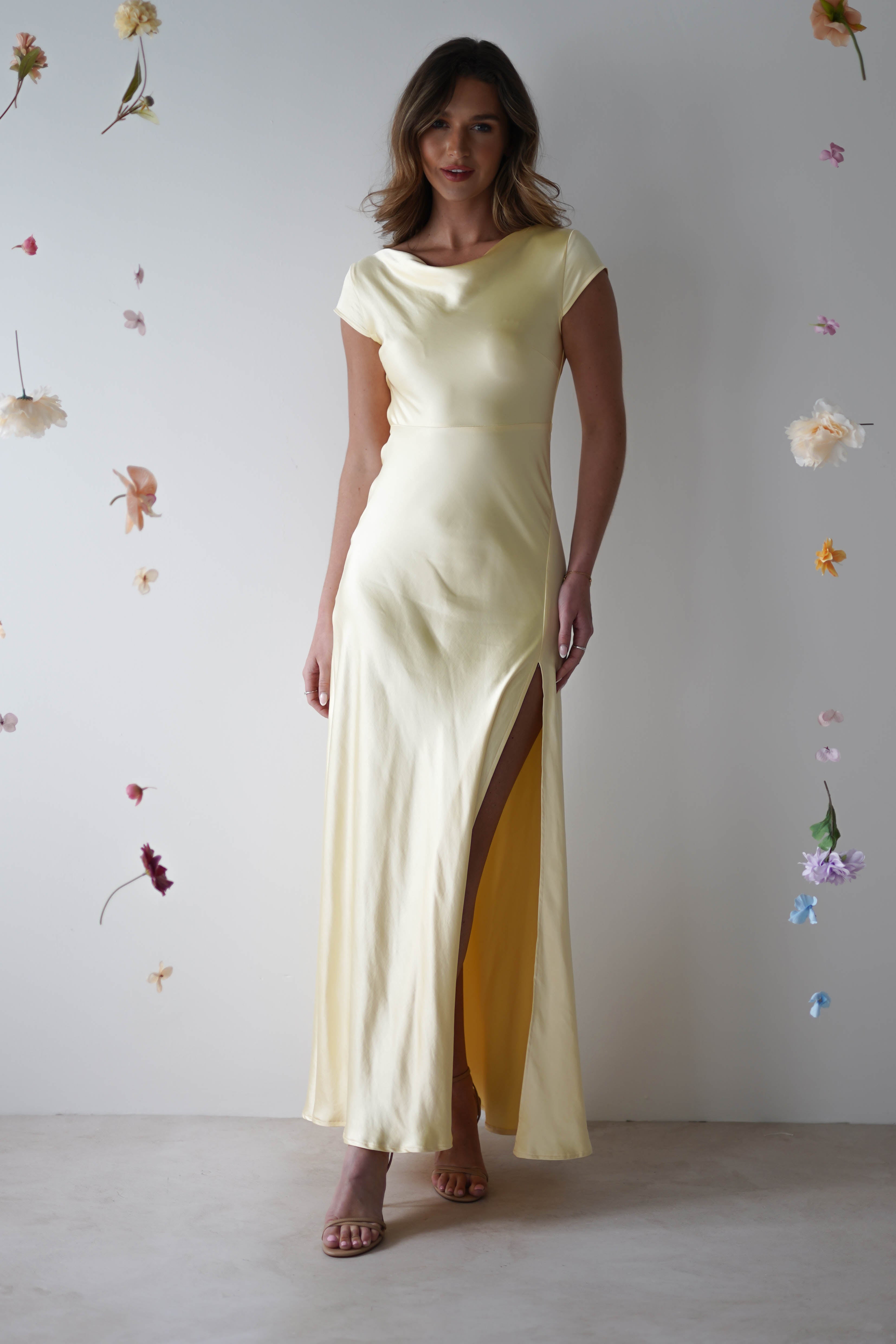 A woman wearing a pale yellow maxi slip dress with short sleeves and a thigh-high slit stands against a plain wall decorated with hanging pastel flowers—perfect for bridesmaids or special occasions.