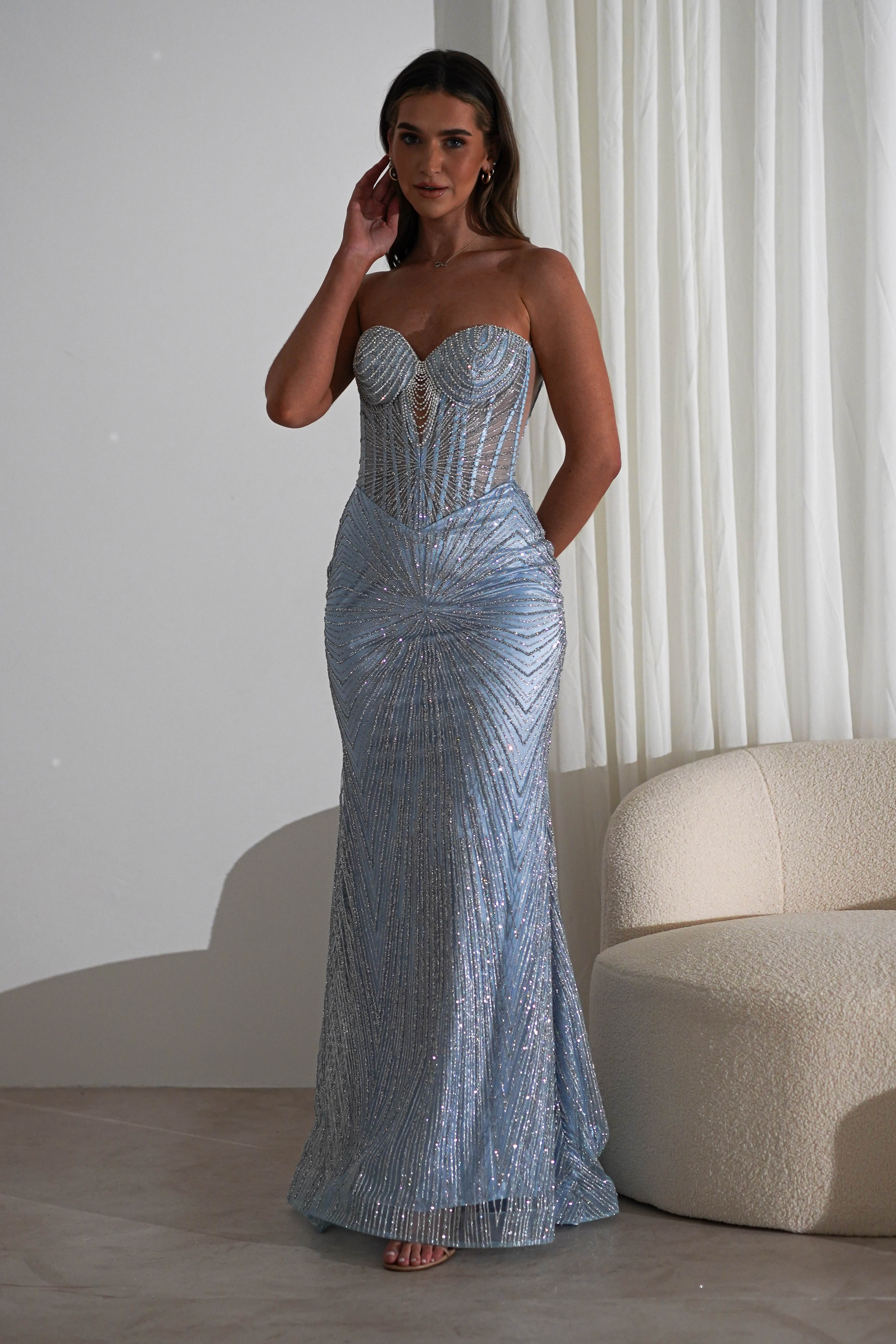 A woman in a strapless, shimmering blue embellished gown with intricate patterns stands in a softly lit room. The background features white curtains and a curved beige chair. She has long hair and poses with one hand touching her face, exuding elegance reminiscent of a timeless prom dress.