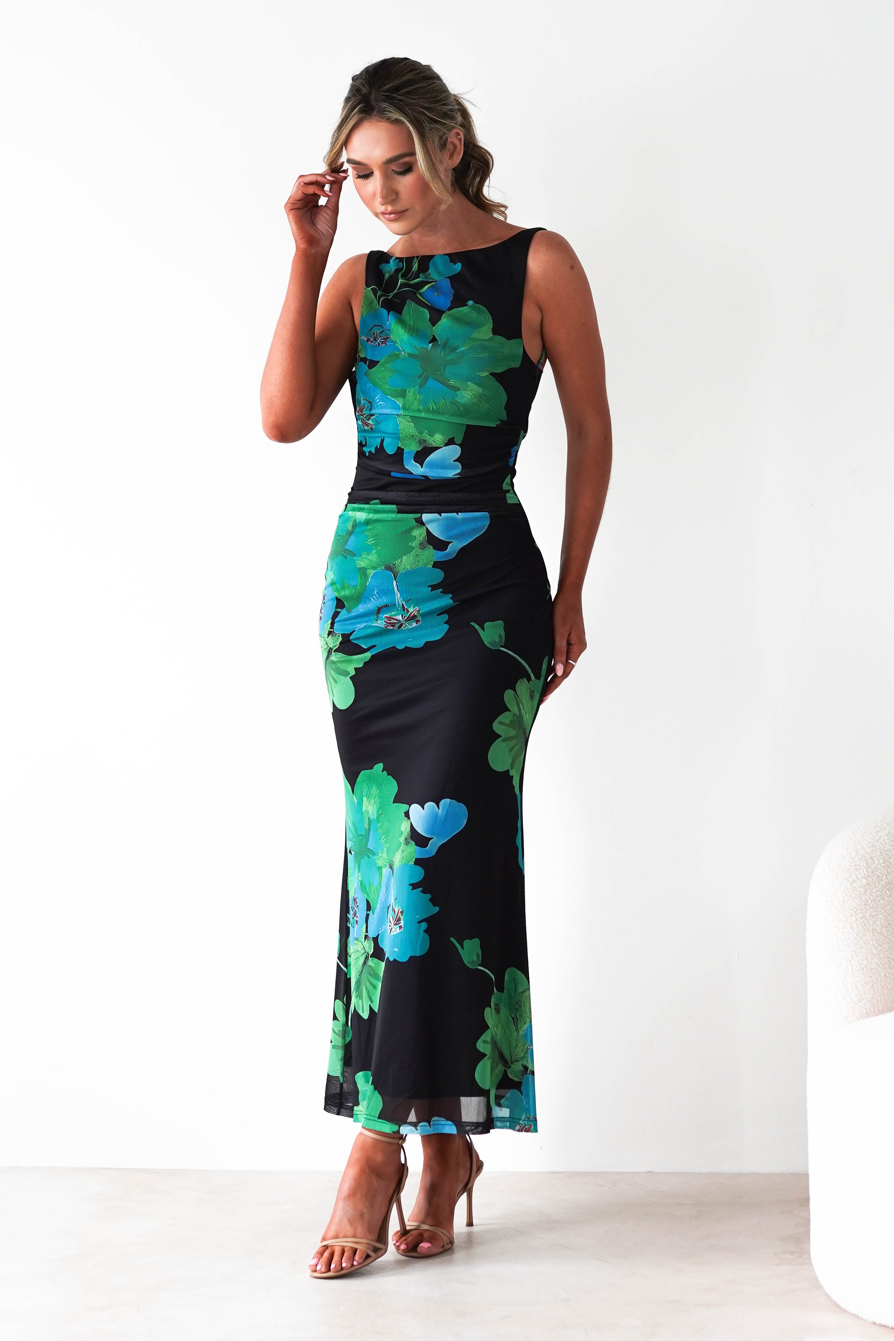 Woman wearing a sleeveless black maxi dress with bold green and blue tropical floral prints, standing against a white background with hands on hips.