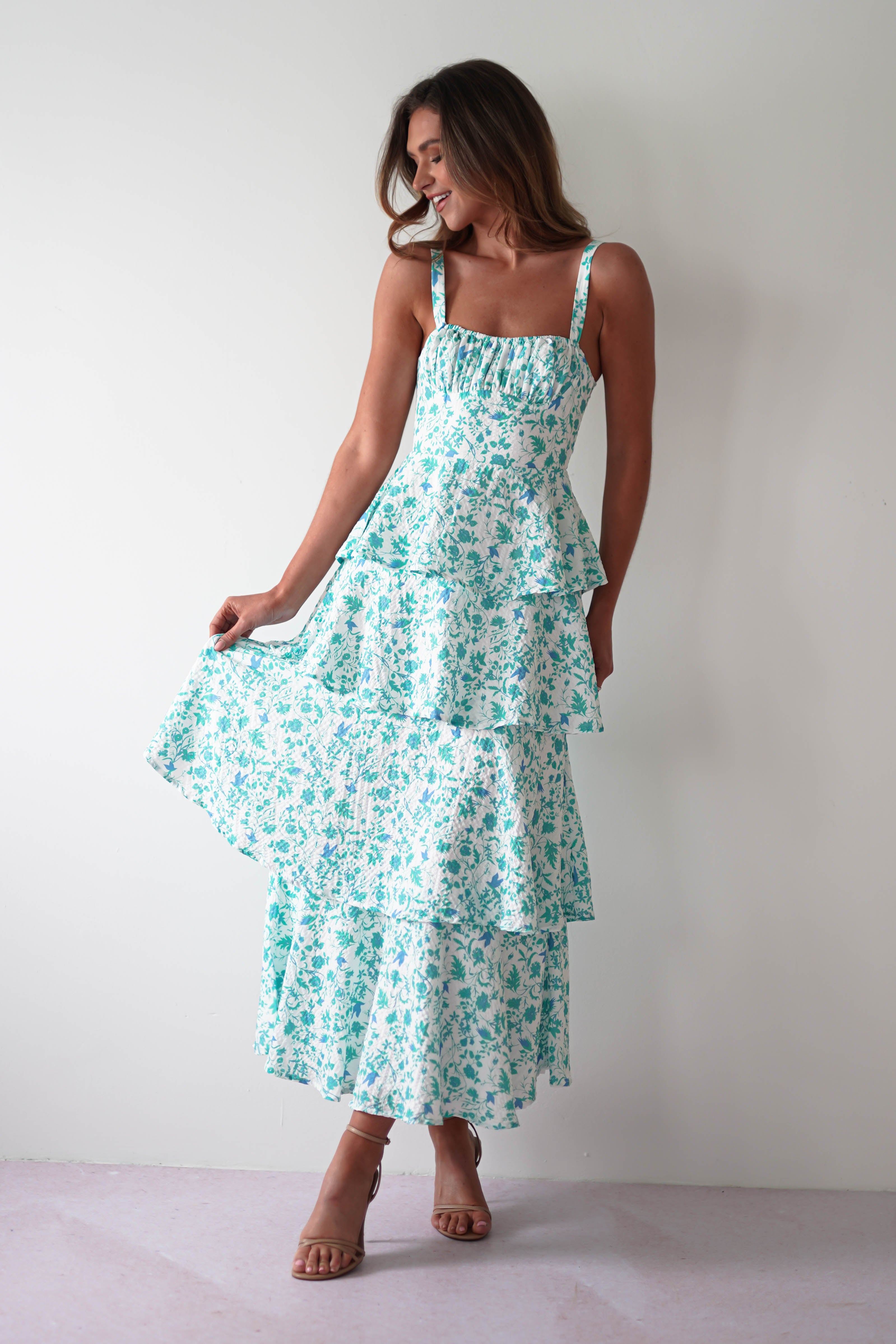 A person wearing a long, tiered summer print maxi dress with shades of turquoise and white stands gracefully against a plain background. Beautiful shoulder ties accent the dress as they hold part of it, showcasing its flow and pattern. They are also wearing sandals.