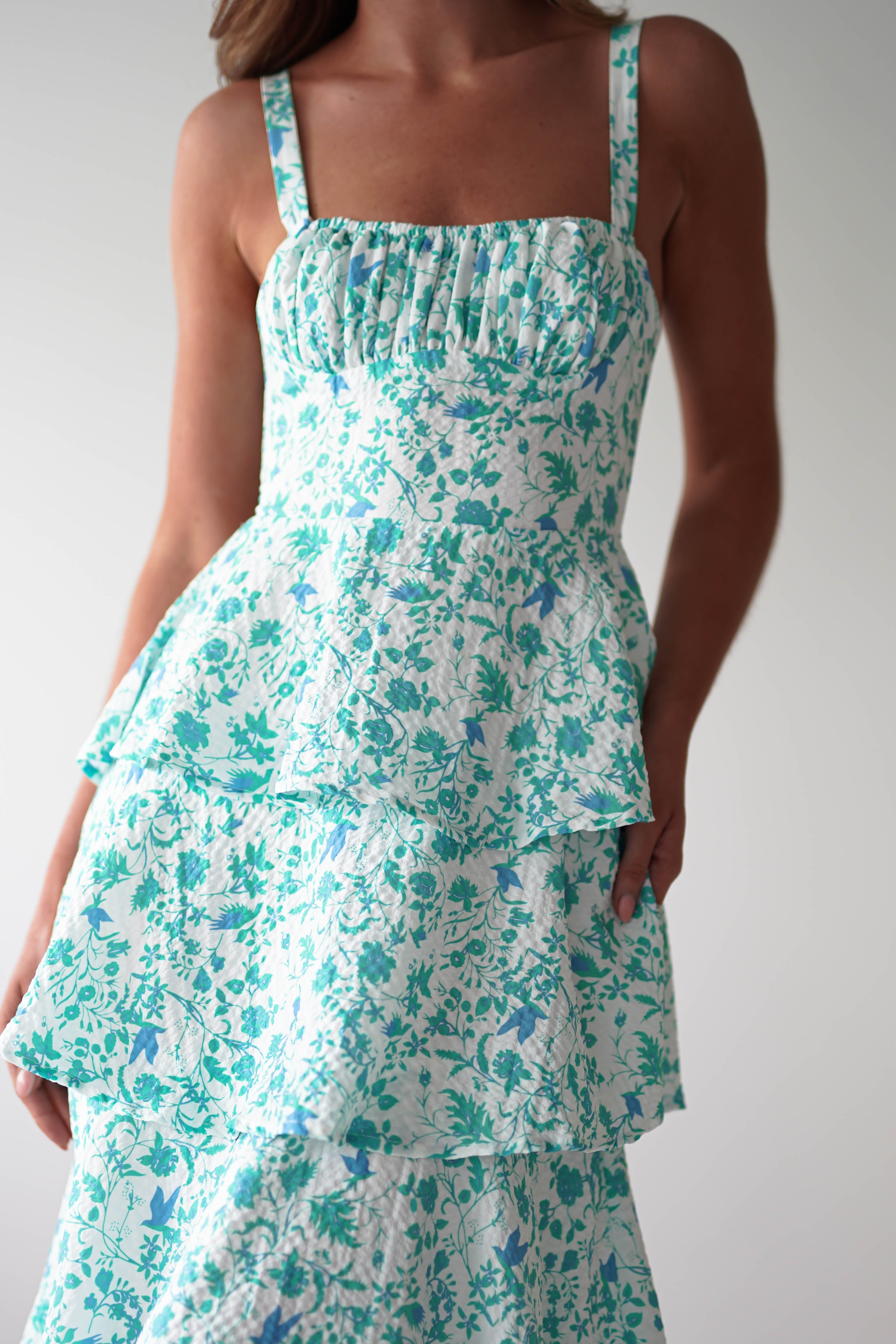 A woman wearing a sleeveless, tiered floral maxi dress with blue and white patterns. The summer print features a ruched bodice and a flowing skirt. Beautiful shoulder ties add charm, while the plain, light-colored background highlights her style—perfect for wedding guests.