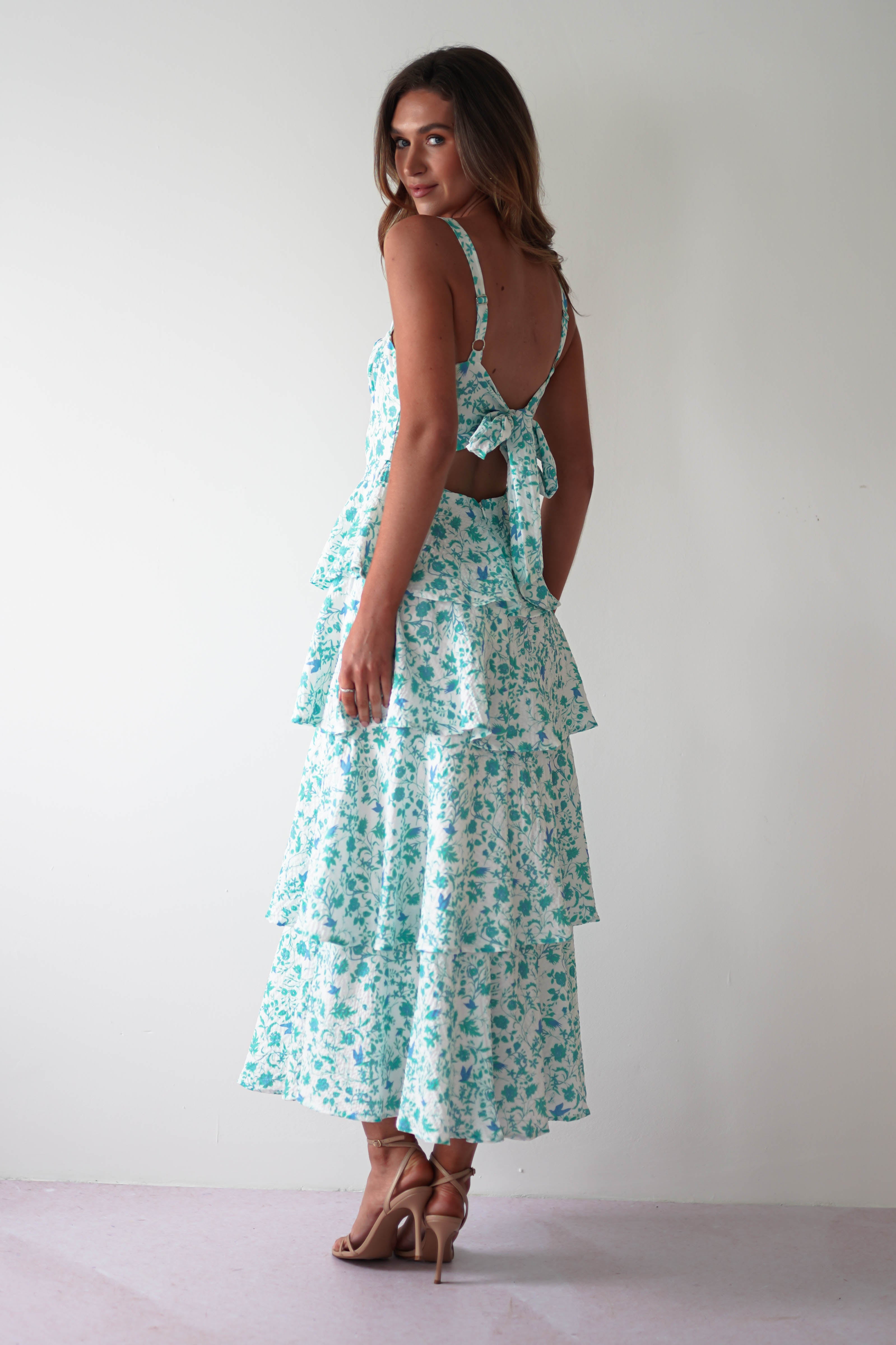 A woman in a beautiful, flowing blue floral tiered dress poses against a plain white background. The summer print maxi dress features elegant back and shoulder ties. She is wearing high heels and stands with one hand on her hip, looking over her shoulder—perfect attire for wedding guests.