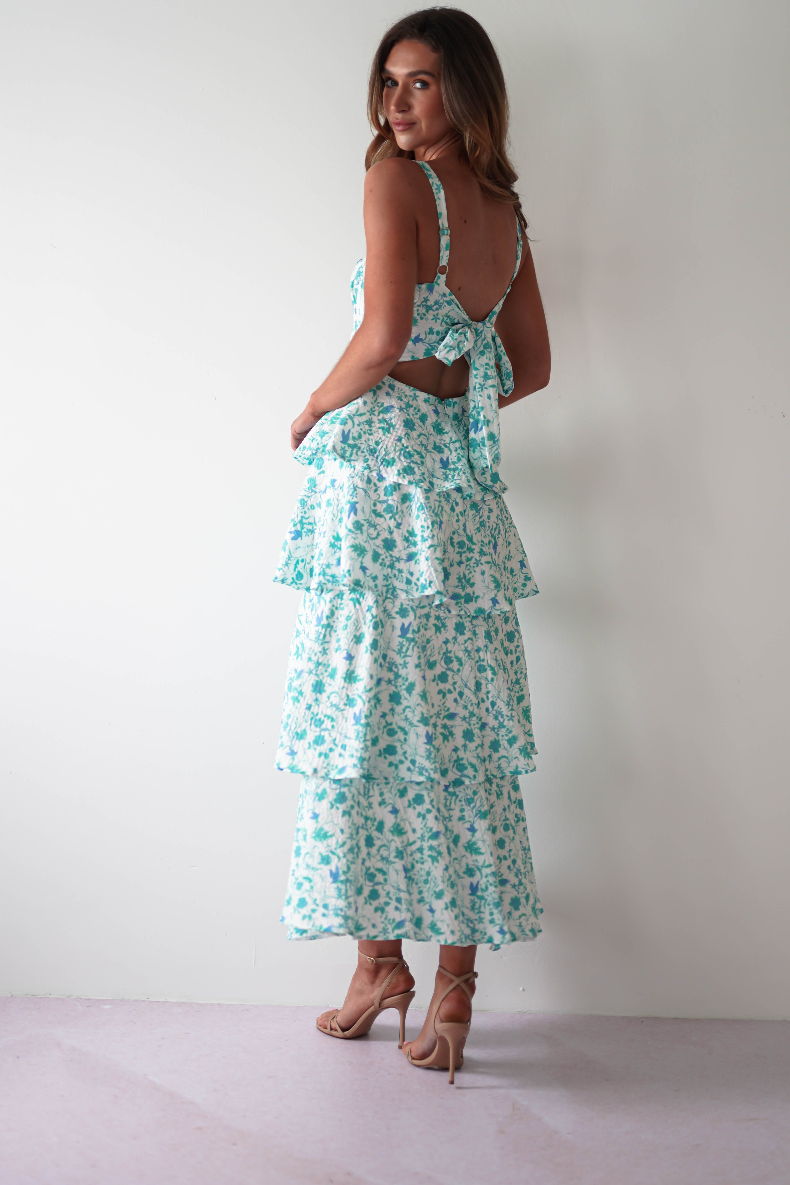 A woman with long hair elegantly glances over her shoulder, showcasing her beautiful shoulder ties. She wears a white and blue floral tiered dress with an open back and strappy heels, perfect for wedding guests. The backdrop is a simple white wall.