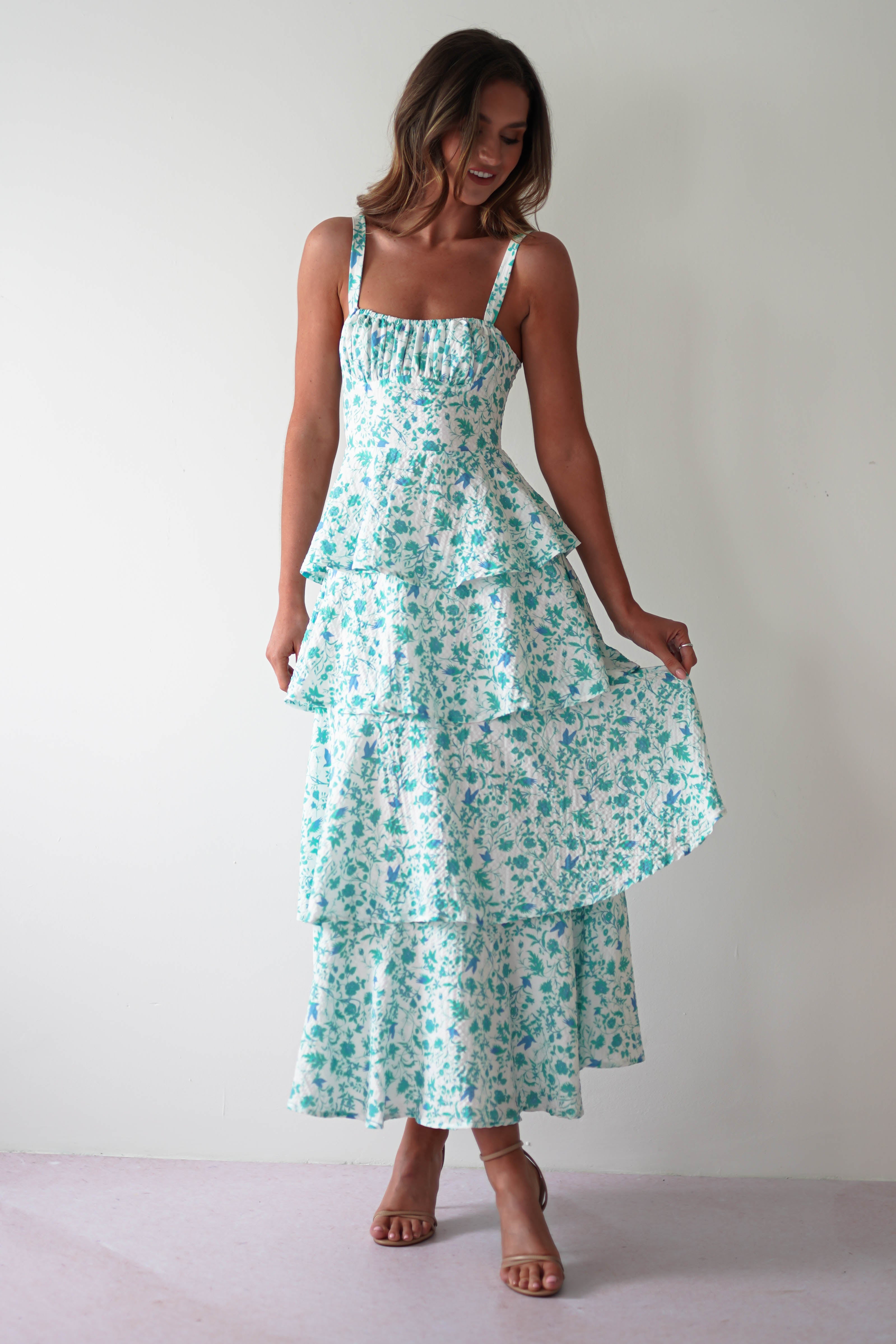 Woman in a sleeveless, tiered white and teal floral dress stands against a plain white background. She holds part of the beautiful summer print maxi dress, smiling and looking down. With tan high heels complementing her elegant look, shes dressed perfectly for any occasion, even as wedding guests.