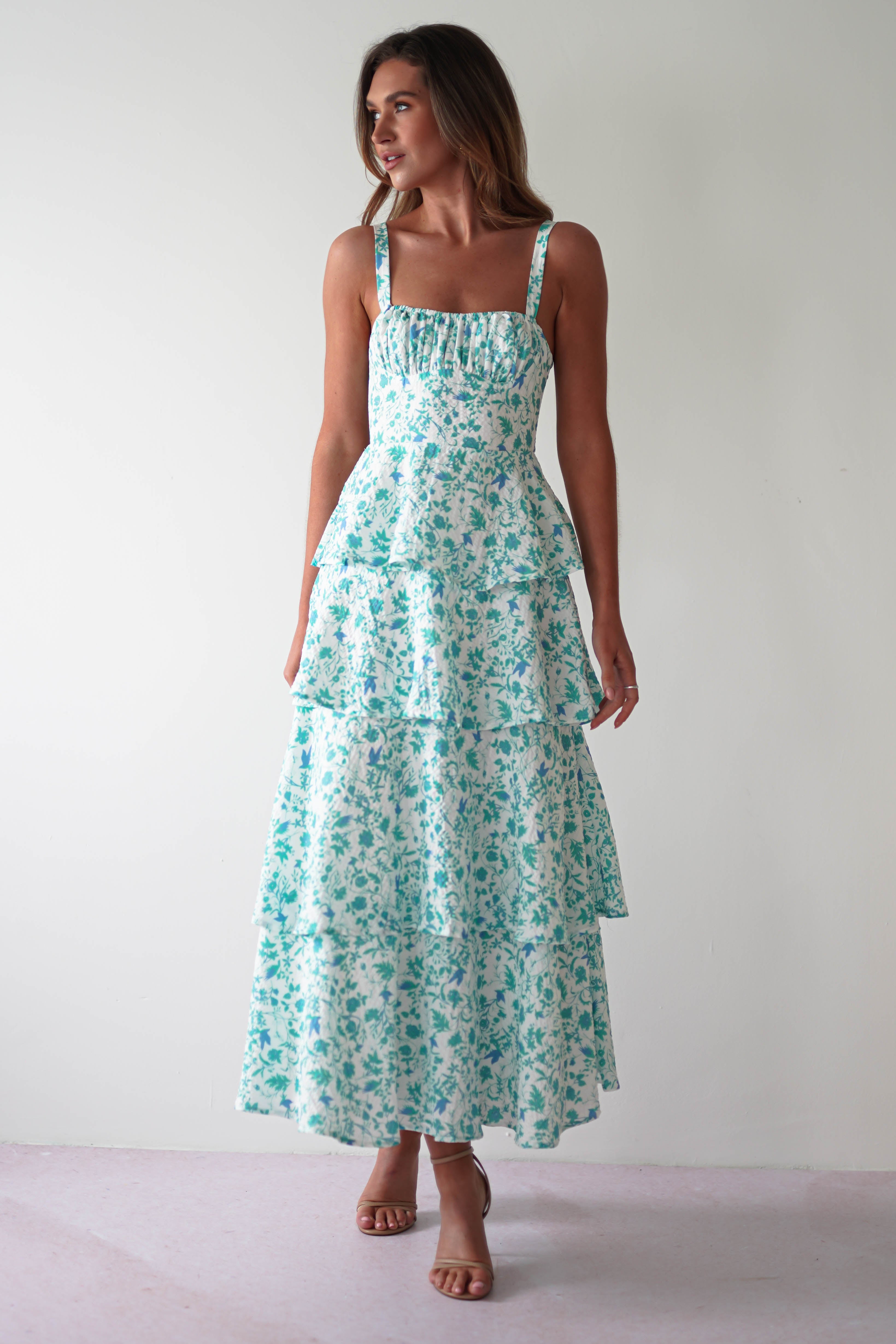 A woman dons a sleeveless, tiered summer print maxi dress adorned with blue floral patterns on a white canvas. With beautiful shoulder ties and neutral-colored high heels, she stands gracefully to the side, an elegant choice for wedding guests.