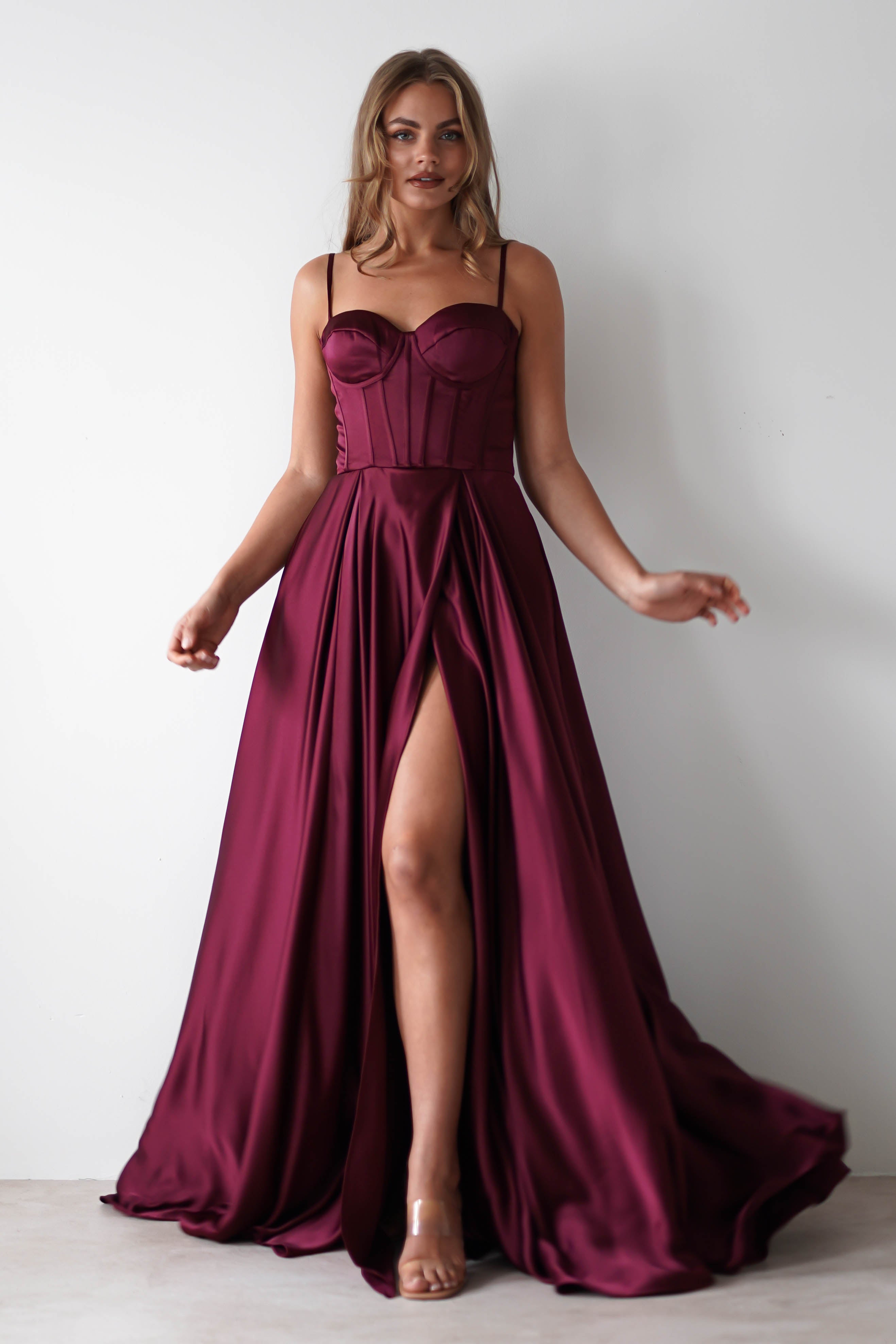 A woman in a sleeveless, deep burgundy satin gown with a corset-style bodice, lace-up back, and high front slit poses against a plain white background. She wears clear high heels and has loose, wavy hair—perfect for formal events.