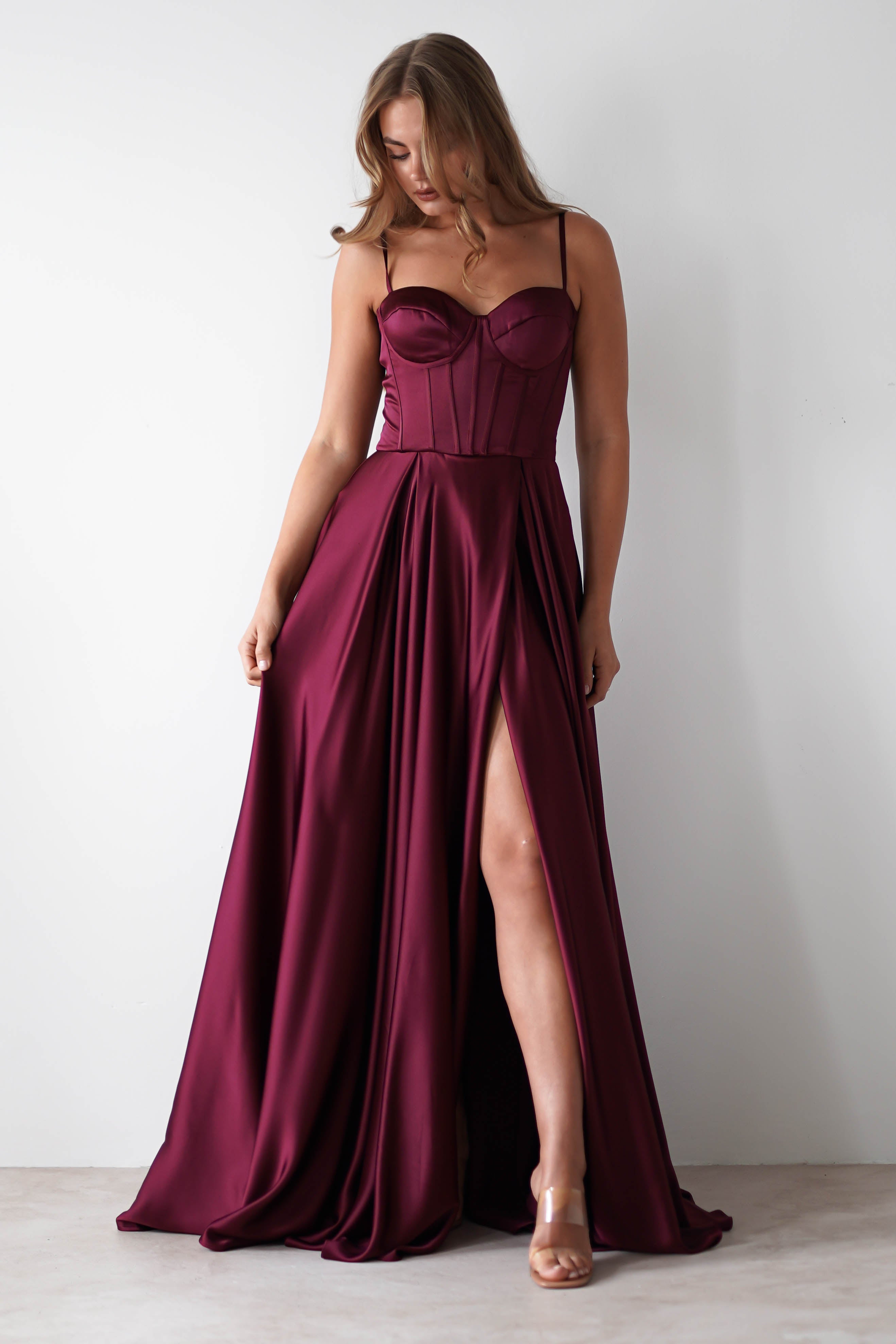 A woman wears a deep burgundy satin gown with a lace-up back, corset-style bodice, thin straps, and a high slit—perfect for formal events. She stands against a plain white wall, looking down.